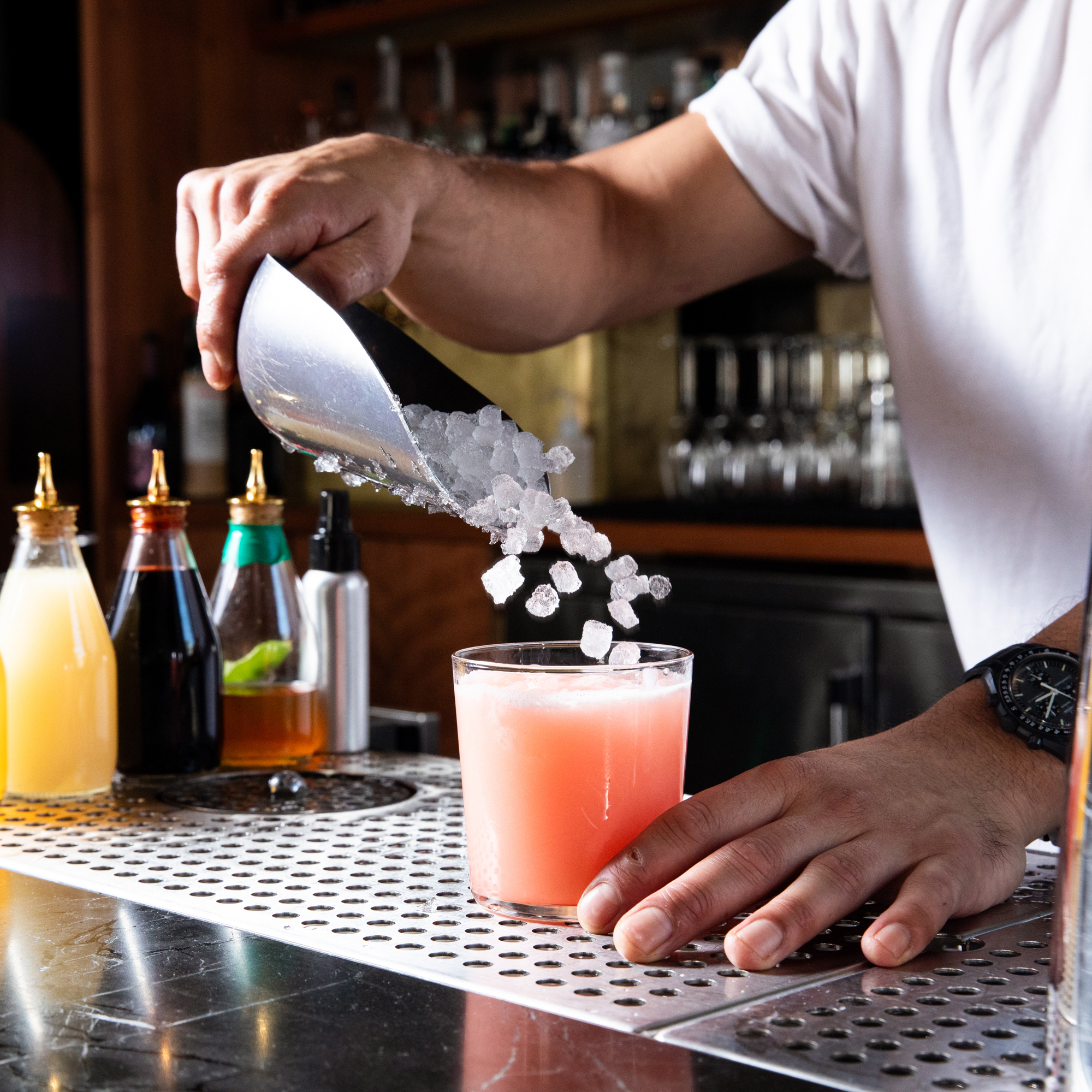 A bartender pours ice cubes into a pink cocktail at a bar counter, surrounded by various colorful bottled mixers and a metallic counter mat.