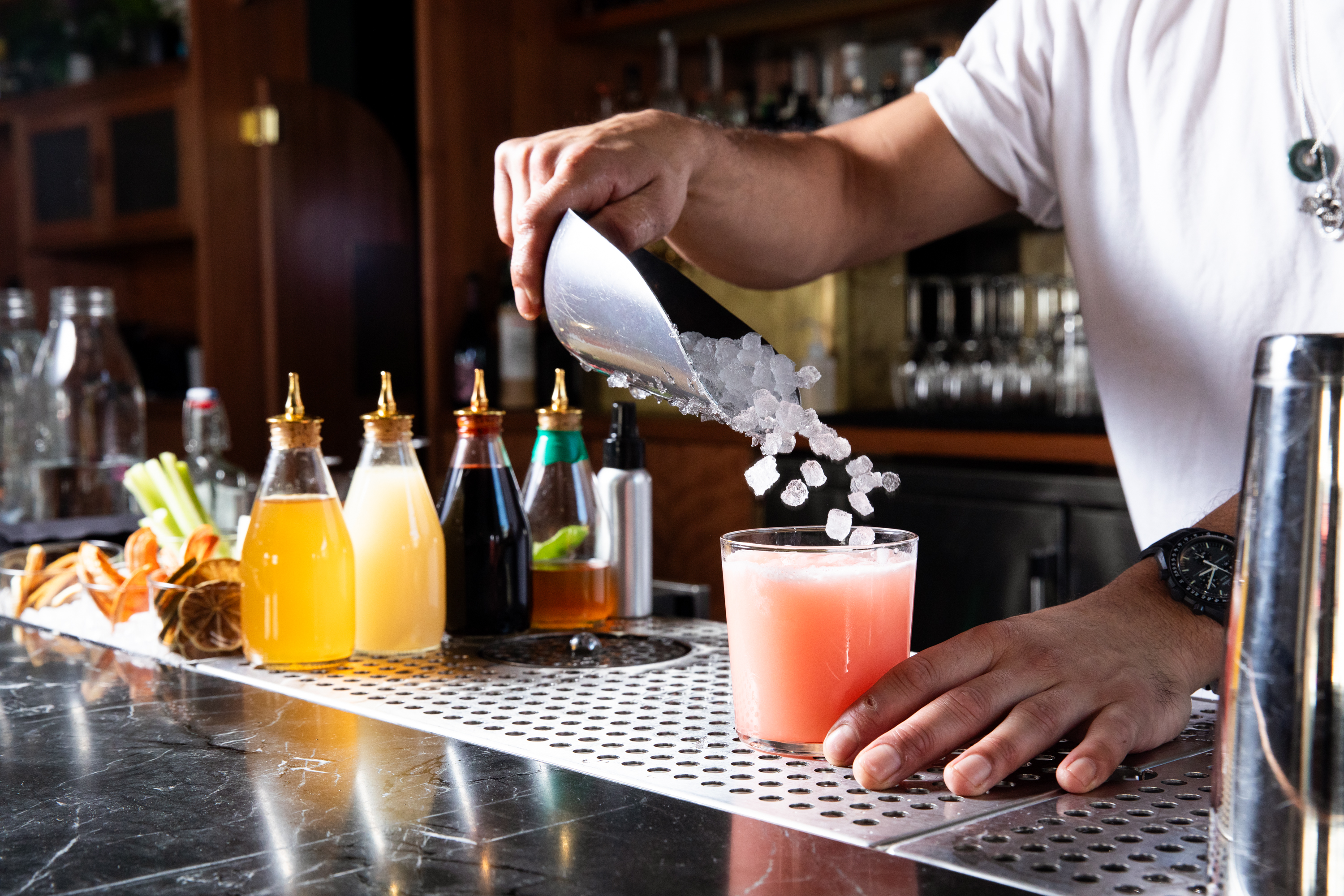 A bartender in a white shirt pours ice into a pink drink using a metal scoop. In the foreground are various syrups and colorful garnishes on the bar.