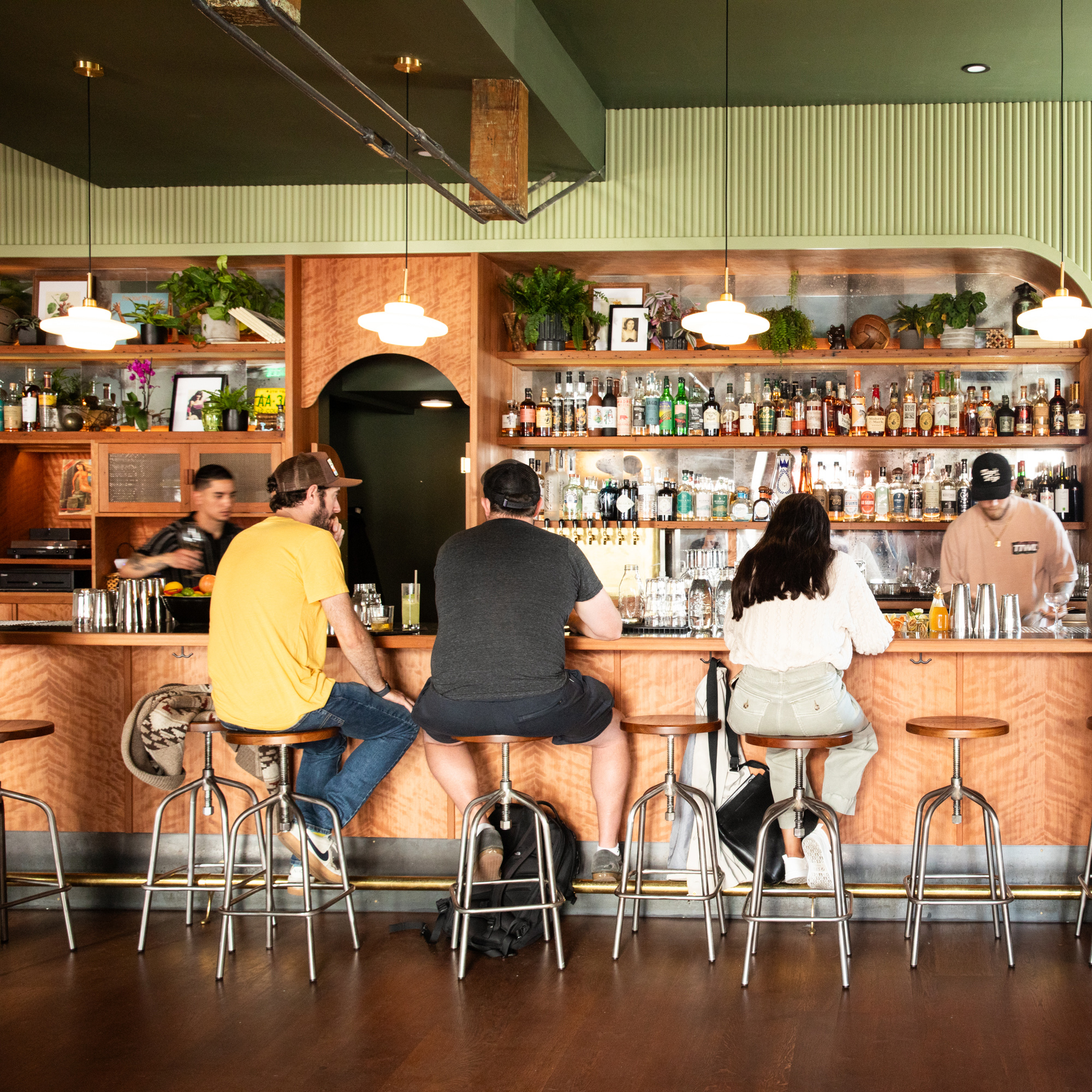 Three people sit at a bar counter surrounded by numerous bottles and plants, while two bartenders prepare drinks. The ambiance is cozy with warm lighting.