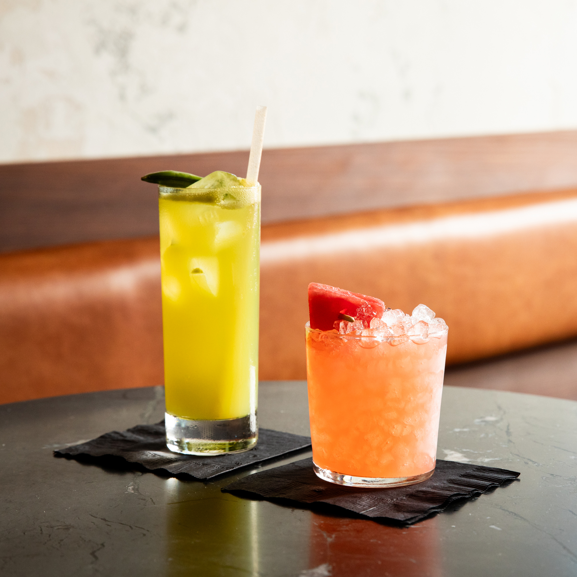 Two cocktails are on a black table with napkins. The left drink is yellow-green in a tall glass with a paper straw and lime garnish. The right drink is pink with crushed ice and a watermelon garnish in a short glass.