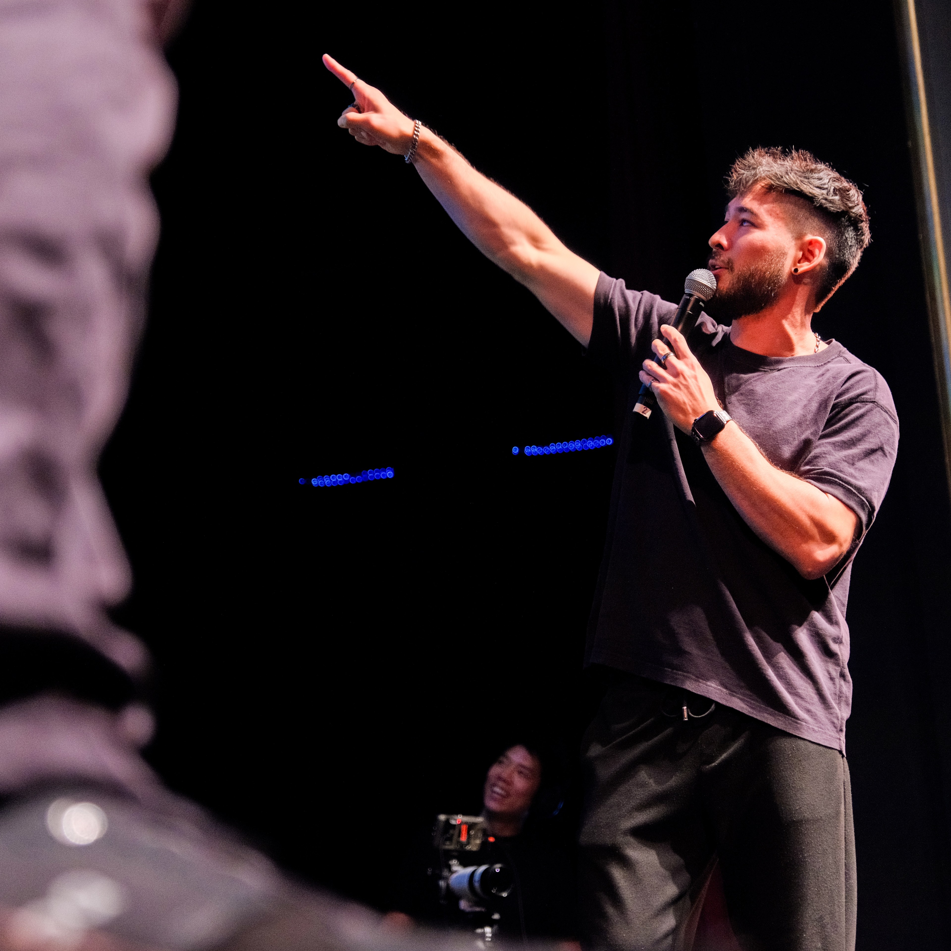 A man with a beard and short hair is holding a microphone and pointing to the sky while speaking on stage, with a camera operator smiling in the background.