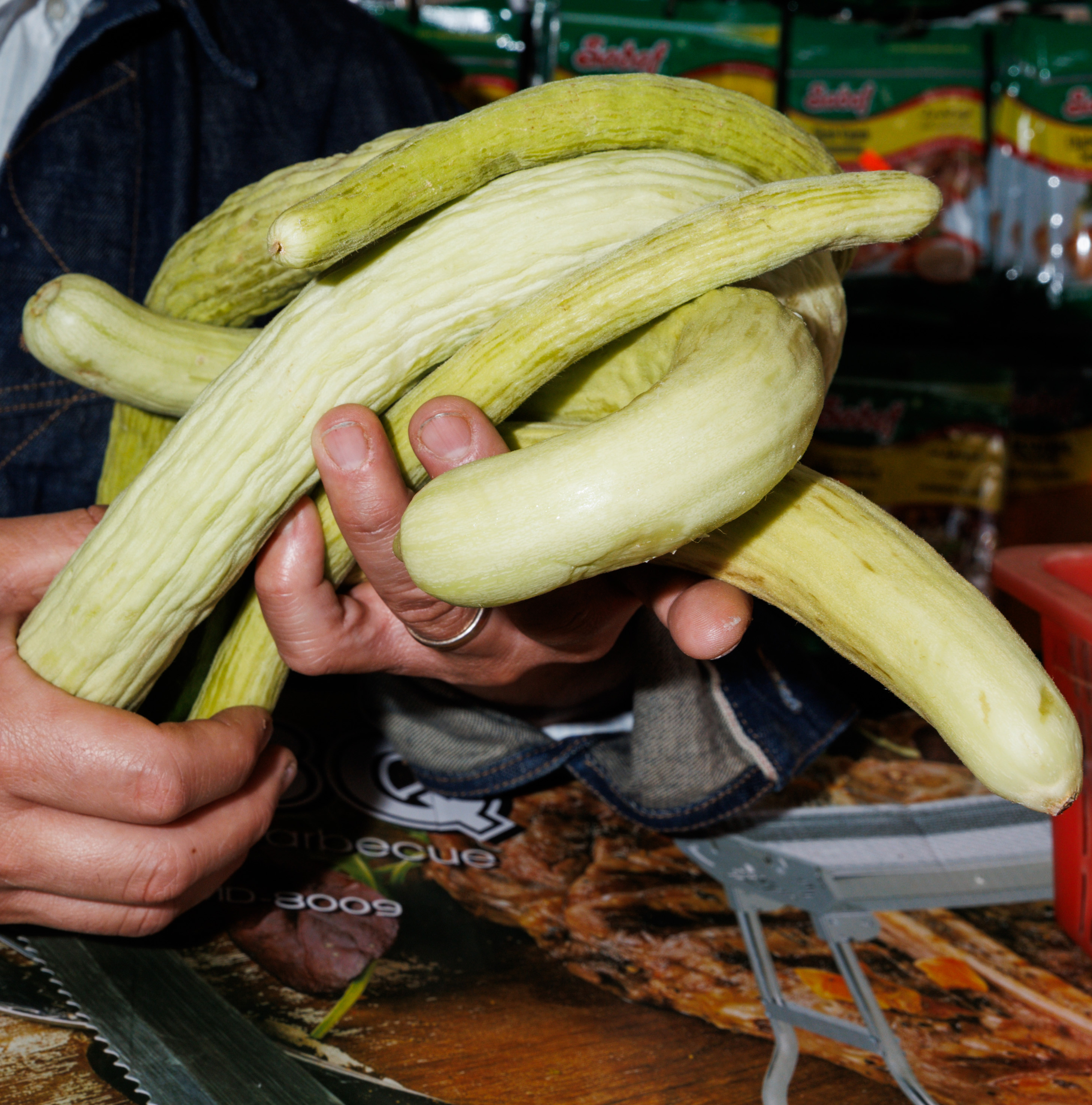 A person is holding several long, yellow-green, curved cucumbers in both hands, with packaged goods visible in the background.