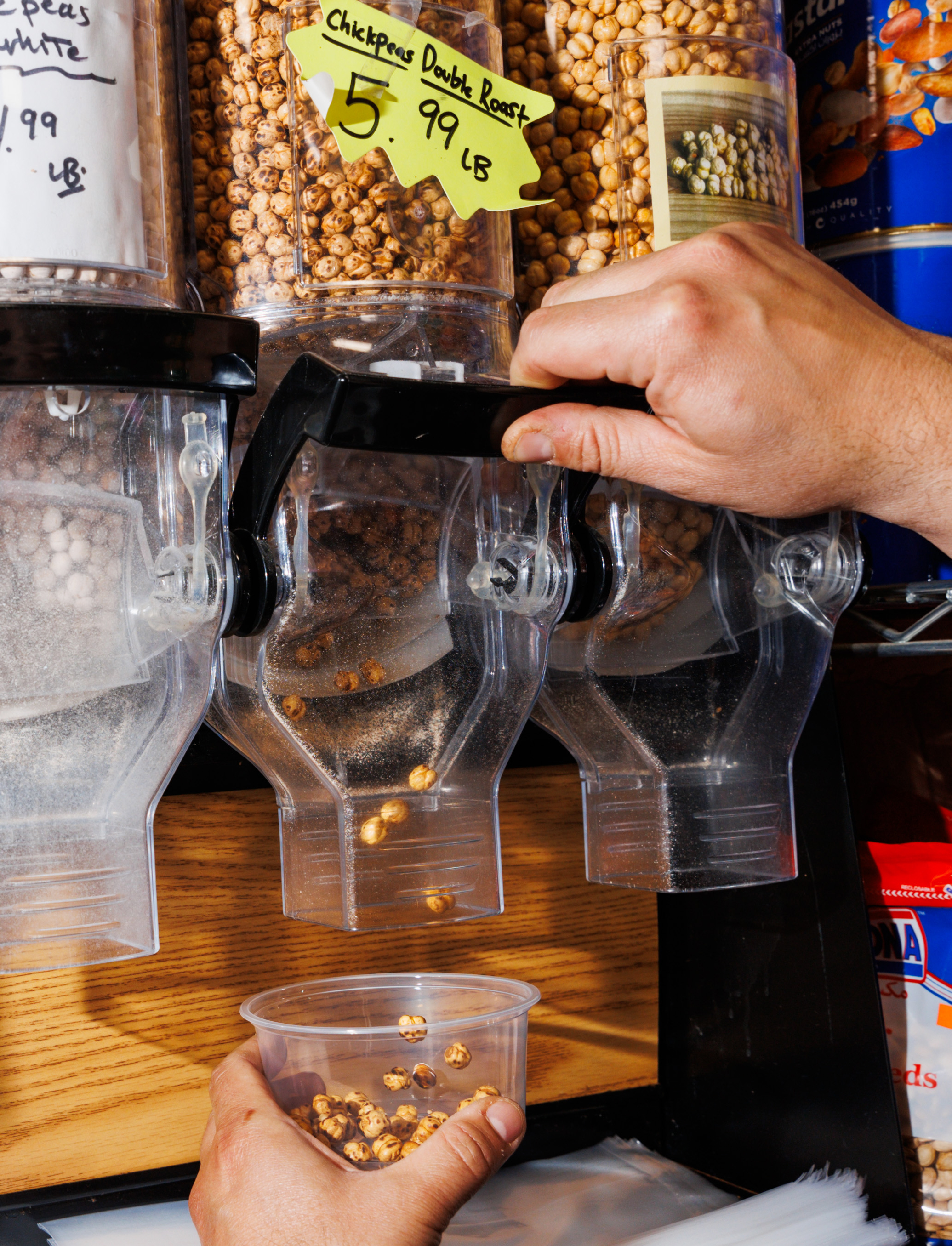 A hand dispenses &quot;Chickpeas Double Roast&quot; from a bulk bin into a plastic container, with a price sign showing $5.99 per pound.