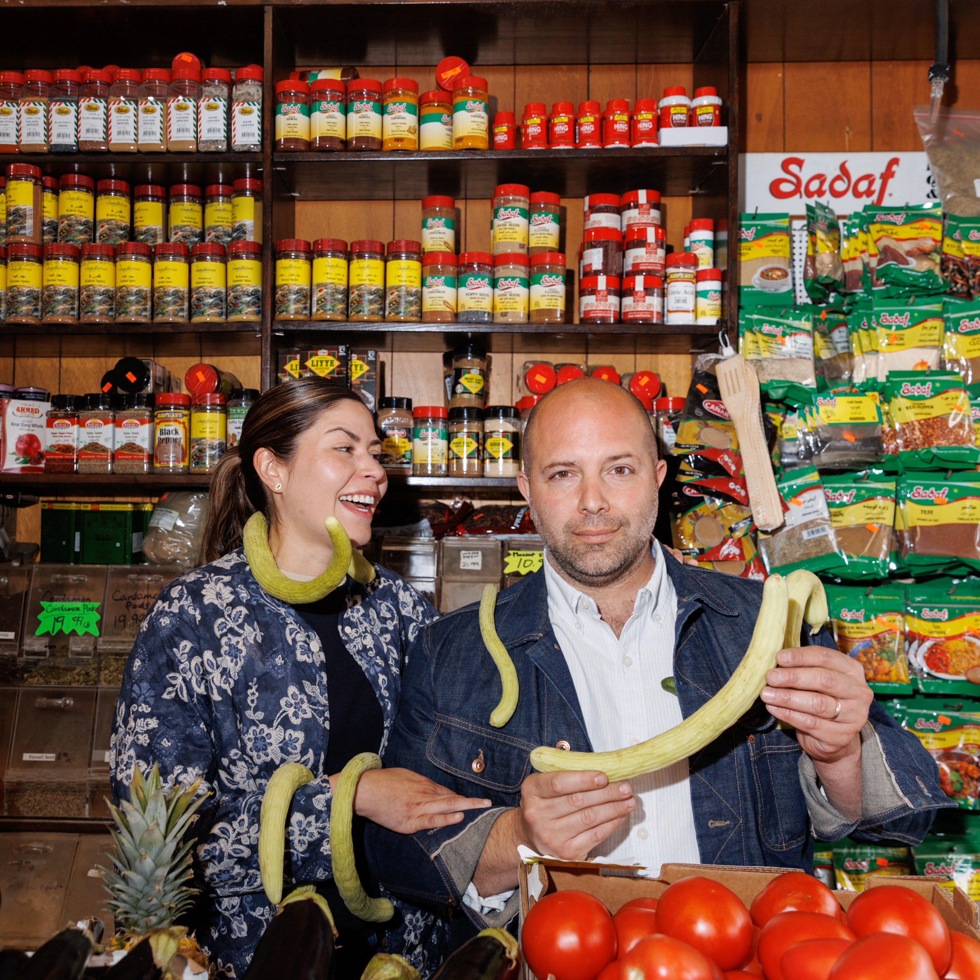 A woman and a bald man stand in a colorful grocery store aisle with spices behind them. The man holds a long vegetable, and both have playful expressions.