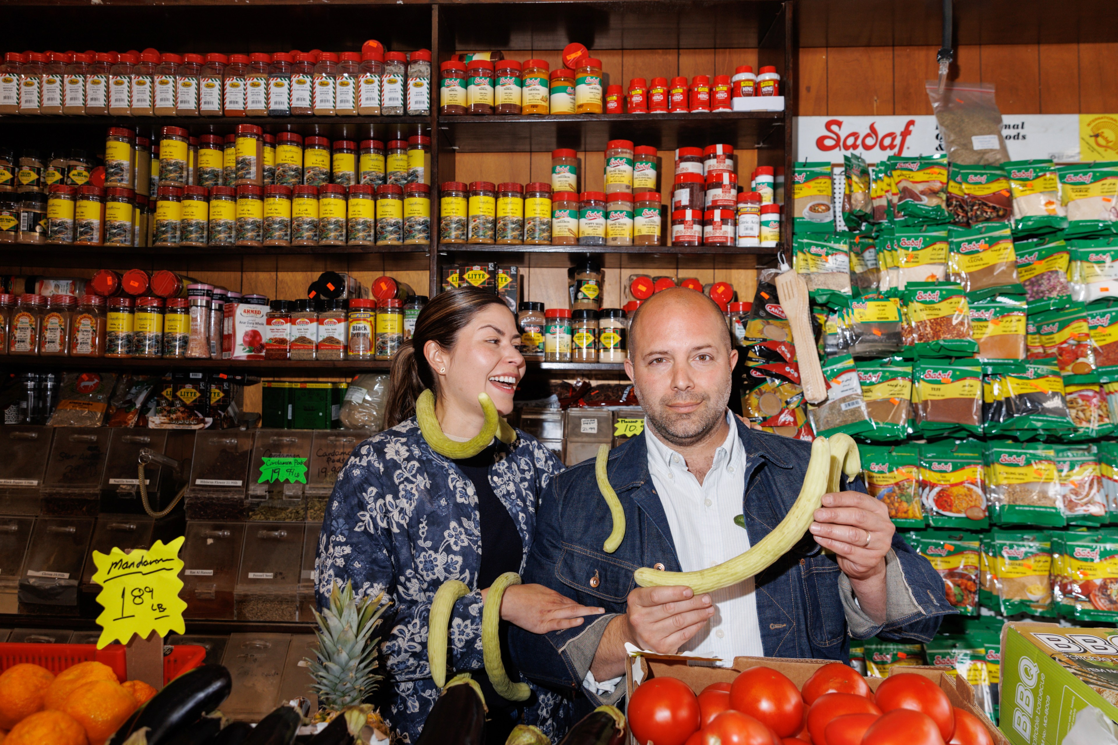 A woman and a bald man stand in a colorful grocery store aisle with spices behind them. The man holds a long vegetable, and both have playful expressions.