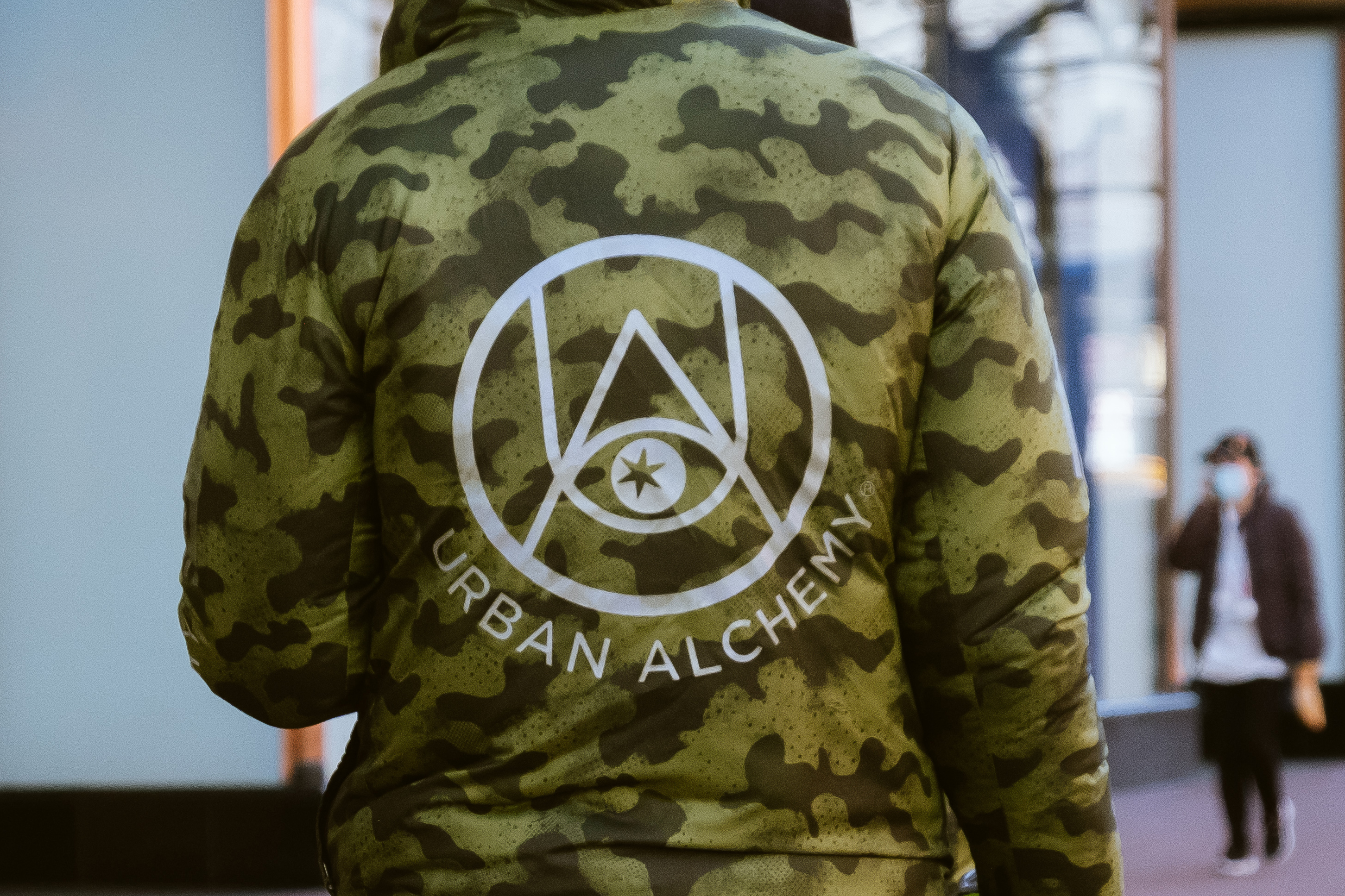 A person is wearing a camouflage jacket with a white circular logo and the words &quot;Urban Alchemy&quot; on the back. Another person is blurred in the background.