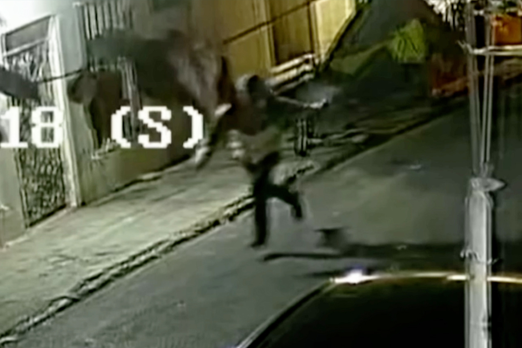 A person is jumping in the air, possibly trying to avoid an object, on a dimly lit street at night, with some buildings and a car in the surroundings.
