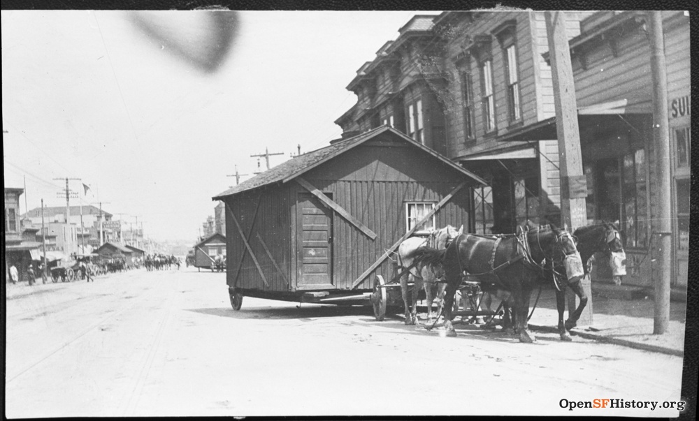 A large shed-like structure is being transported by a horse-drawn cart down a street, with buildings and power lines visible in the background.