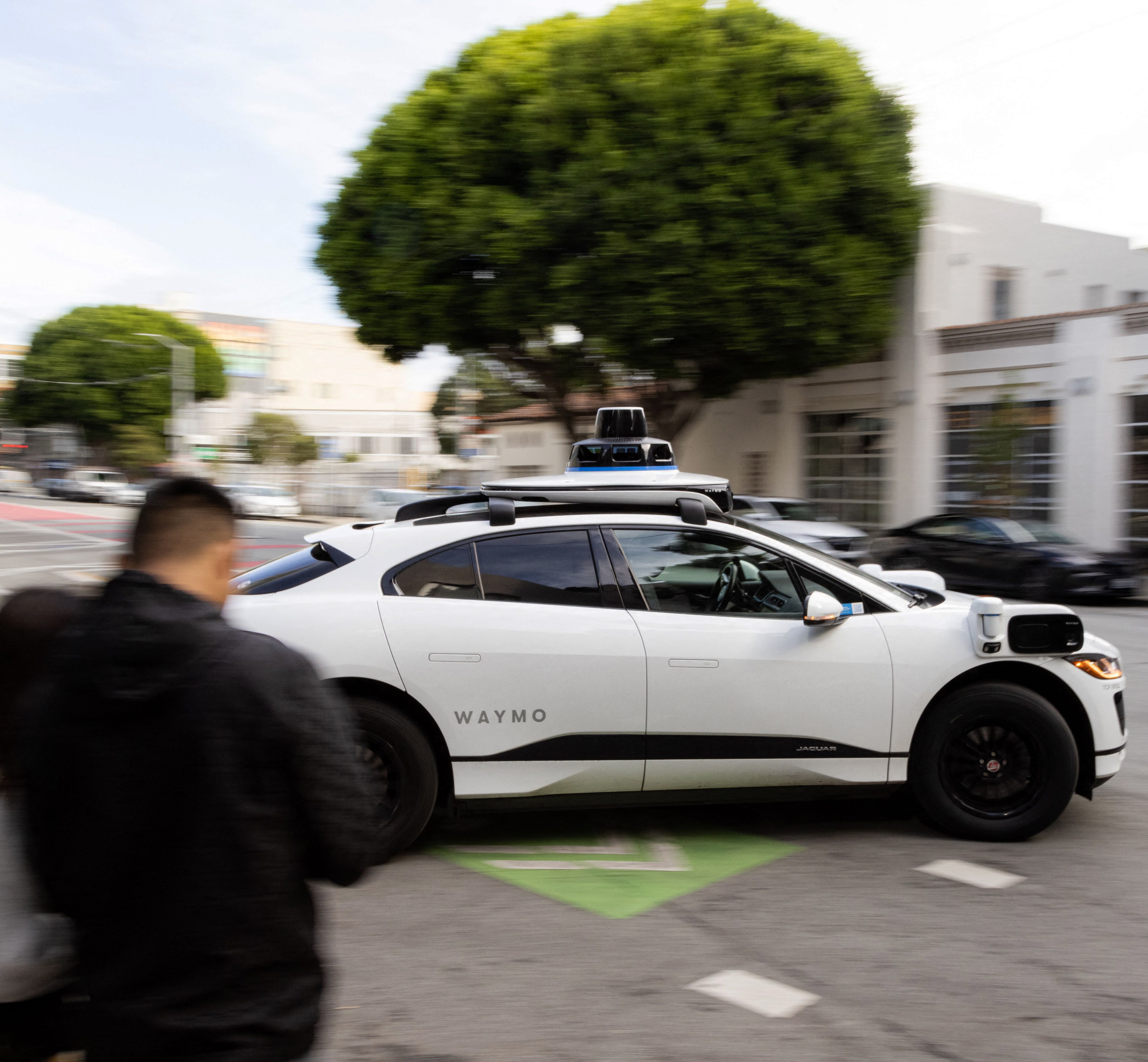 A white Waymo self-driving car, a Jaguar I-PACE, moves through a city street. It has spinning sensors on the roof, and blurry pedestrians are visible in the foreground.