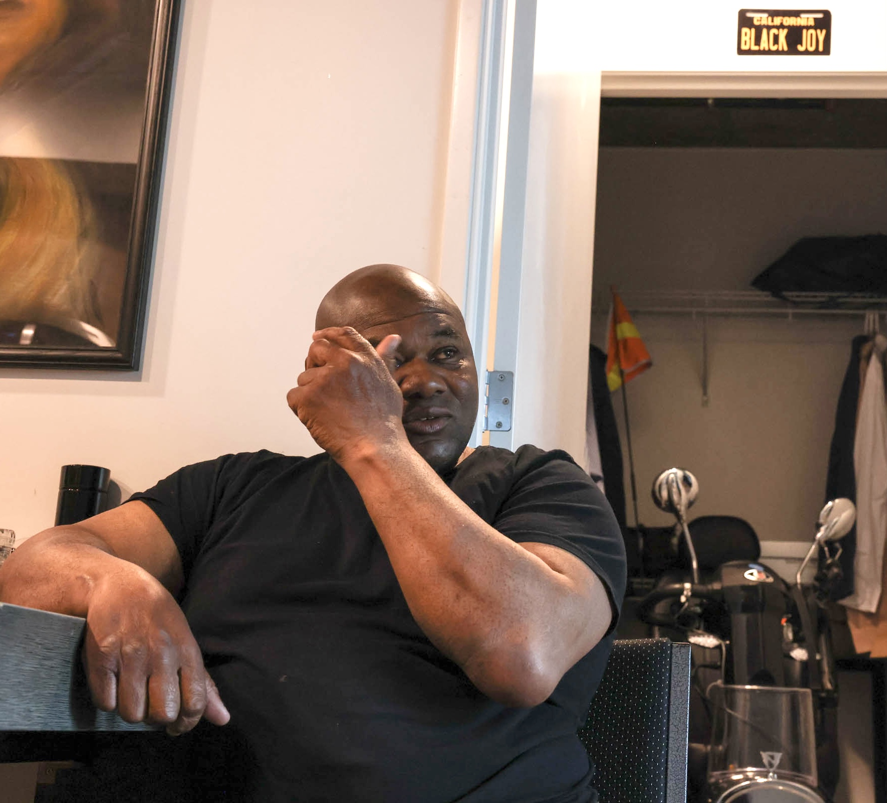 A man in a black shirt sits indoors, leaning on a table and touching his face. Behind him is an open closet with a “BLACK JOY” sign above it.