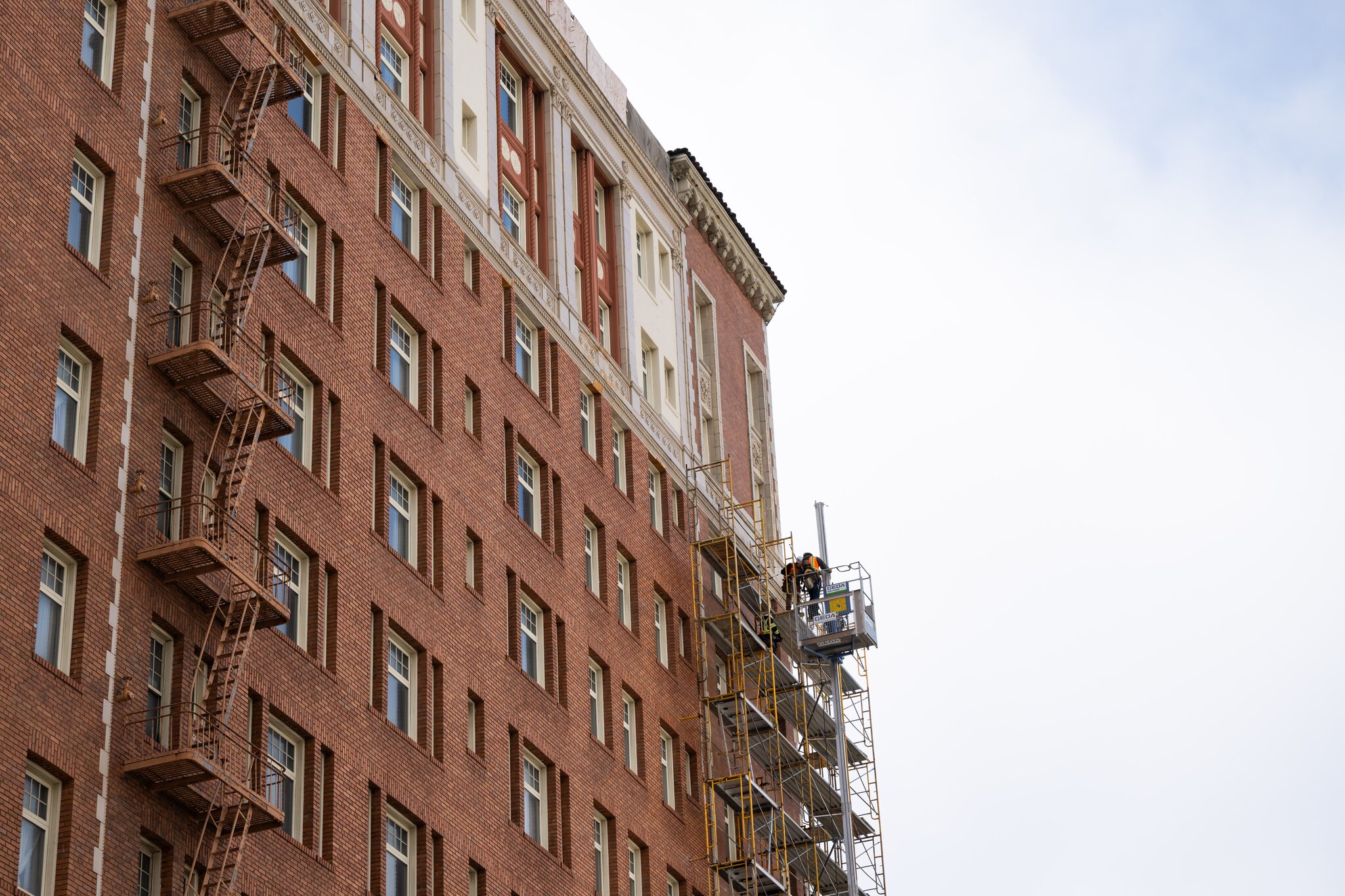 A brick building with symmetrical windows has fire escape stairs on the left side and scaffolding with workers on the right, against a partly cloudy sky.