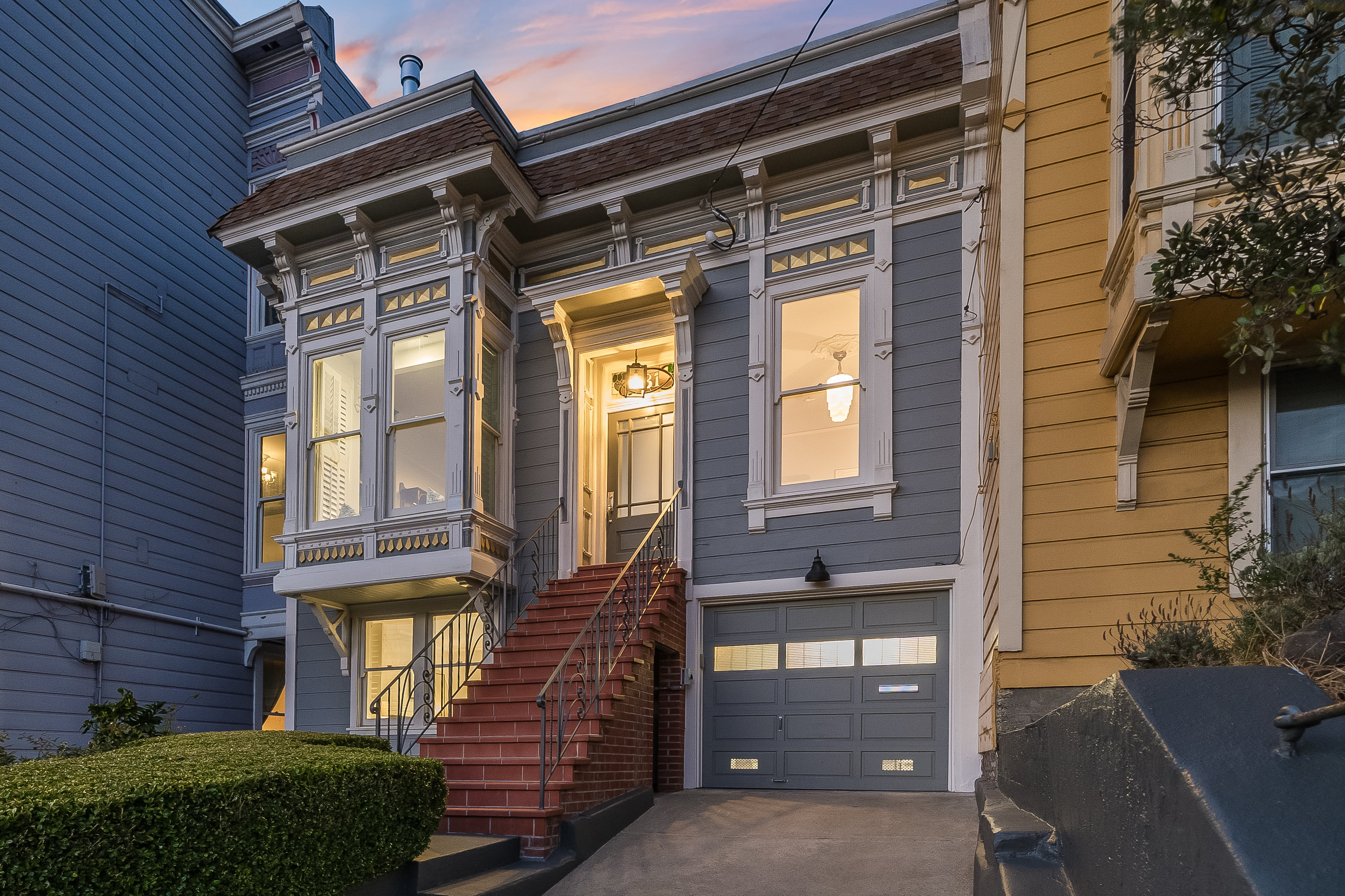 The image shows a charming, two-story house with a light blue exterior, ornate white trim, and large windows; a staircase leads to the front door above a garage.