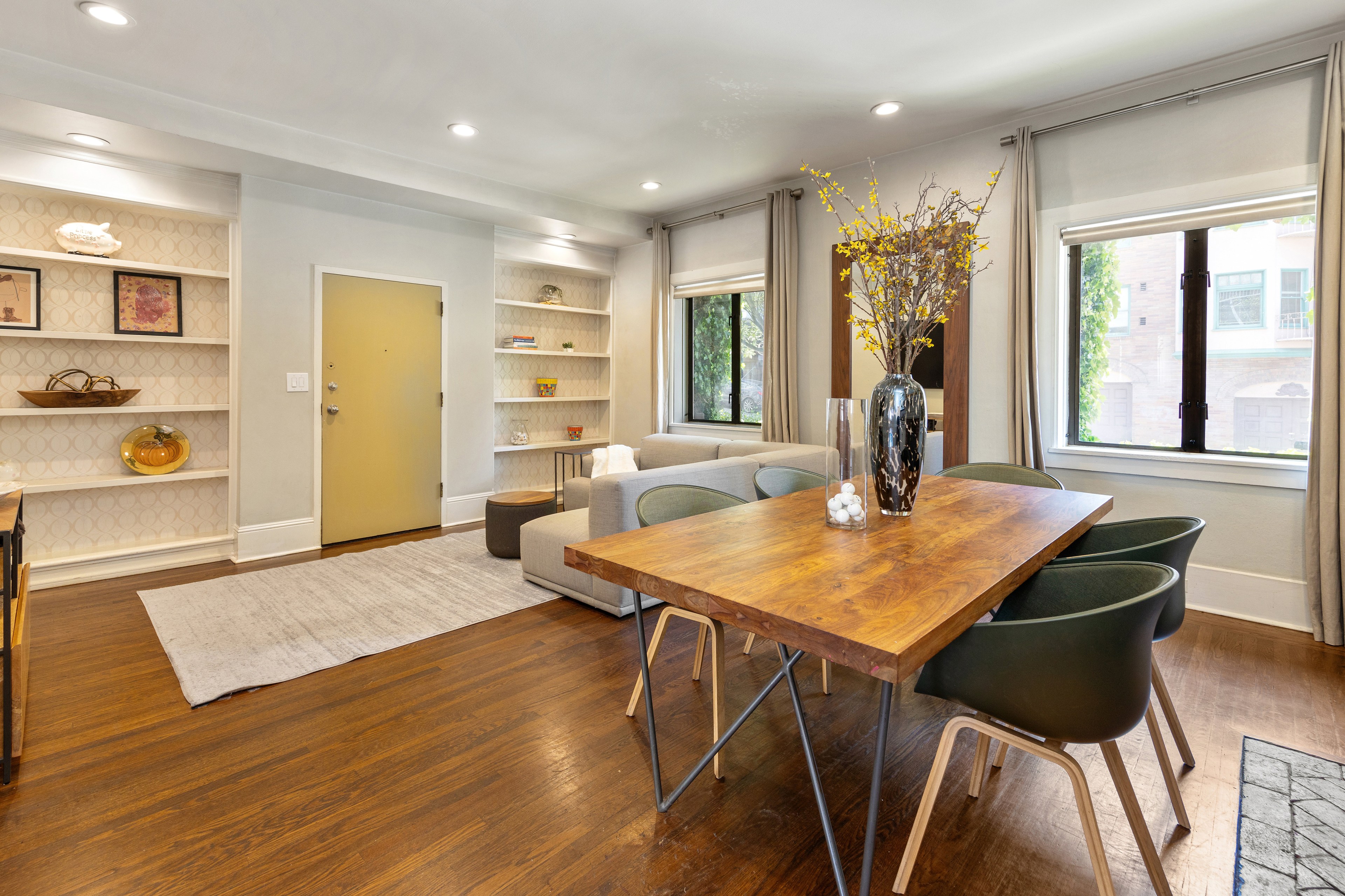 A modern living-dining room features wood floors, a large wooden dining table, gray chairs, built-in shelves, a sofa, and large windows with gray curtains.