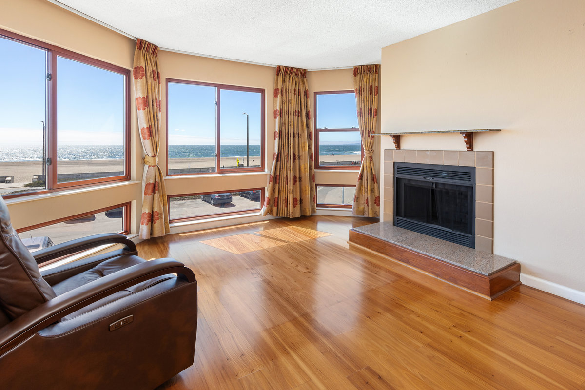 A cozy room with a wooden floor features large windows offering an ocean view, a modern fireplace, and a leather reclining chair, with patterned curtains.