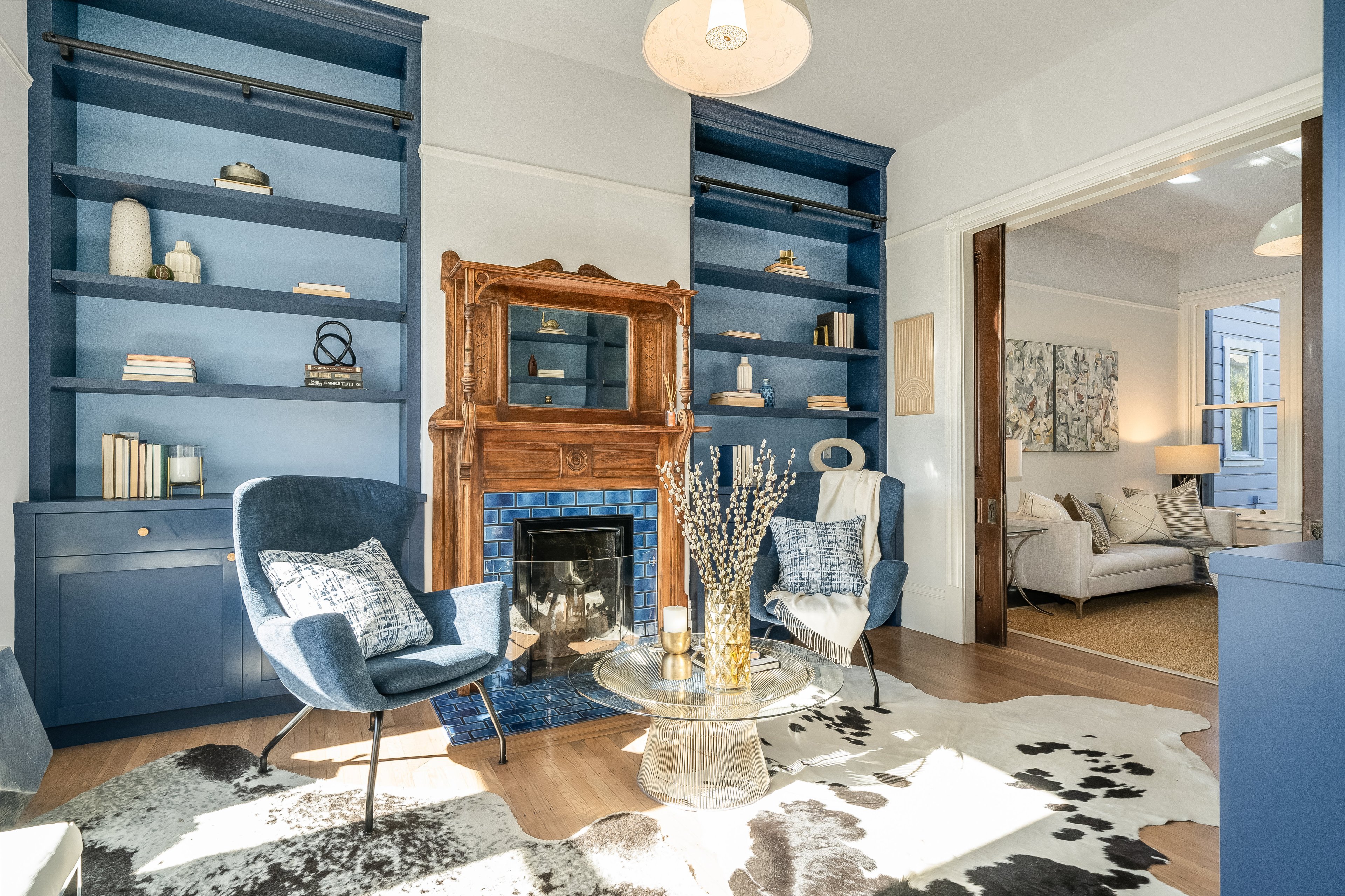 A cozy living room has blue built-in shelving, a wooden mantel, a fireplace, blue chairs, a glass coffee table, and a cowhide rug, leading to another room with a sofa.