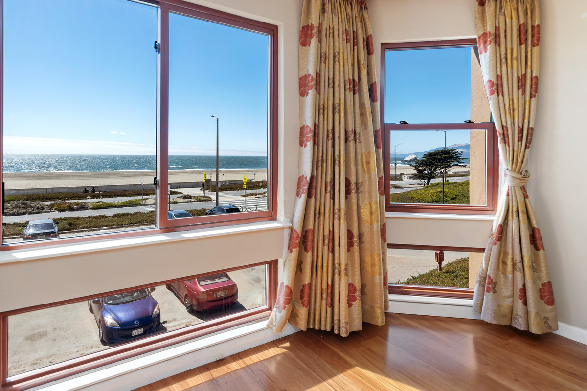 This image shows a scenic ocean view through a bay window adorned with floral-patterned curtains. The window overlooks a roadway and a beach with a bright, clear sky.