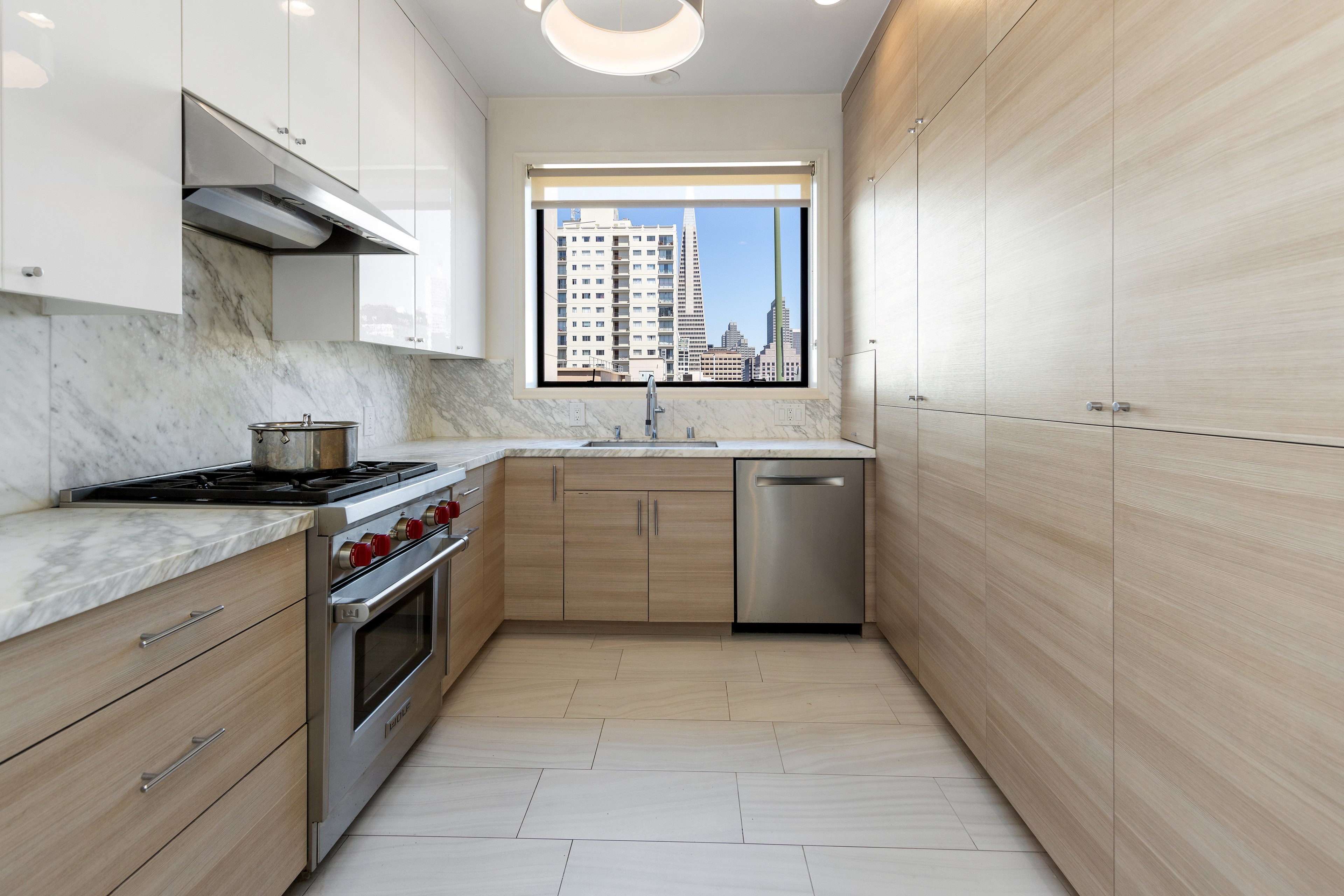 A modern kitchen with light wooden cabinets, marble countertops, gas stove, stainless steel appliances, and a window view of city buildings.