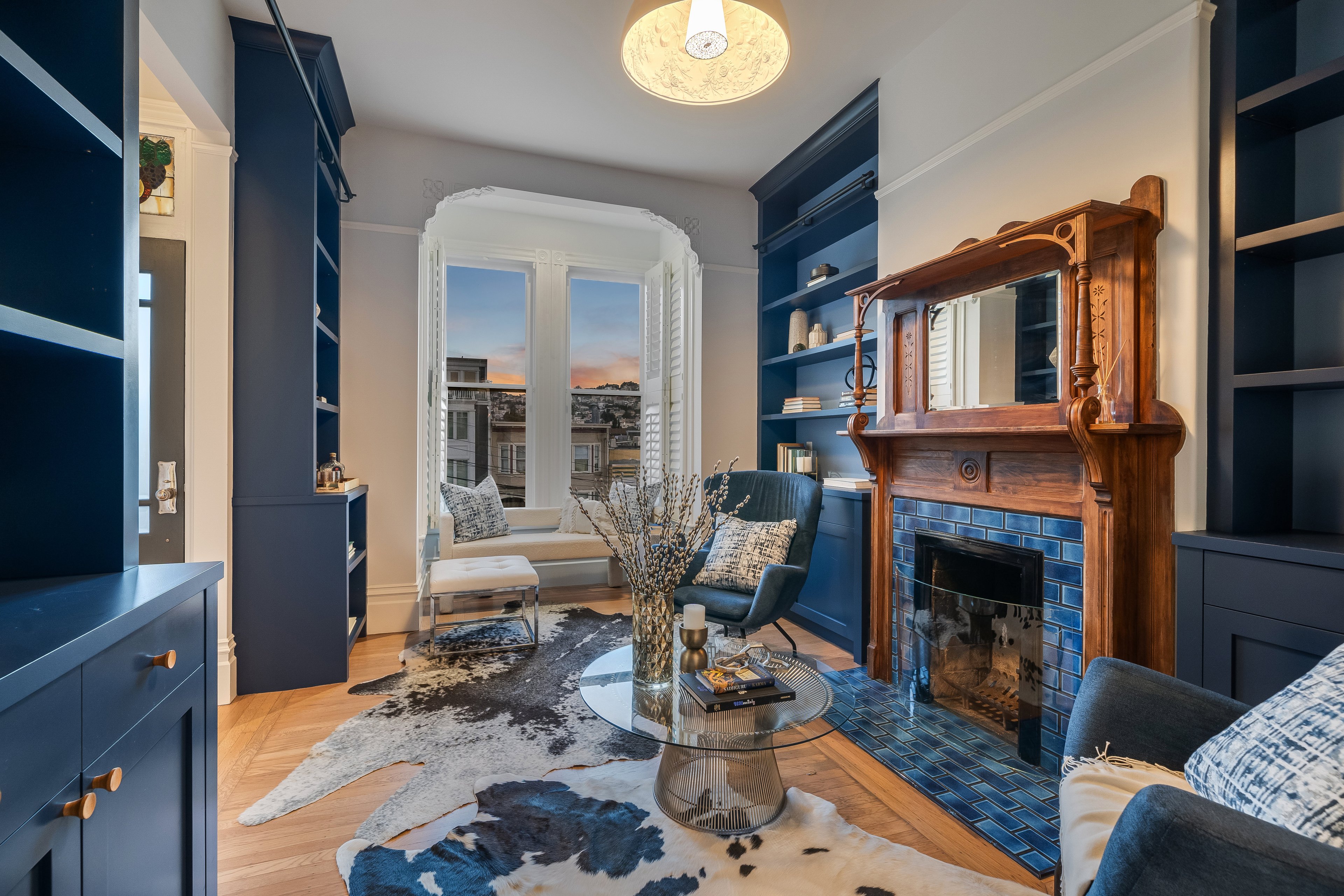 The cozy living room features a wooden fireplace with blue tiles, built-in bookshelves, armchairs, a glass coffee table, and large windows showcasing a cityscape view.