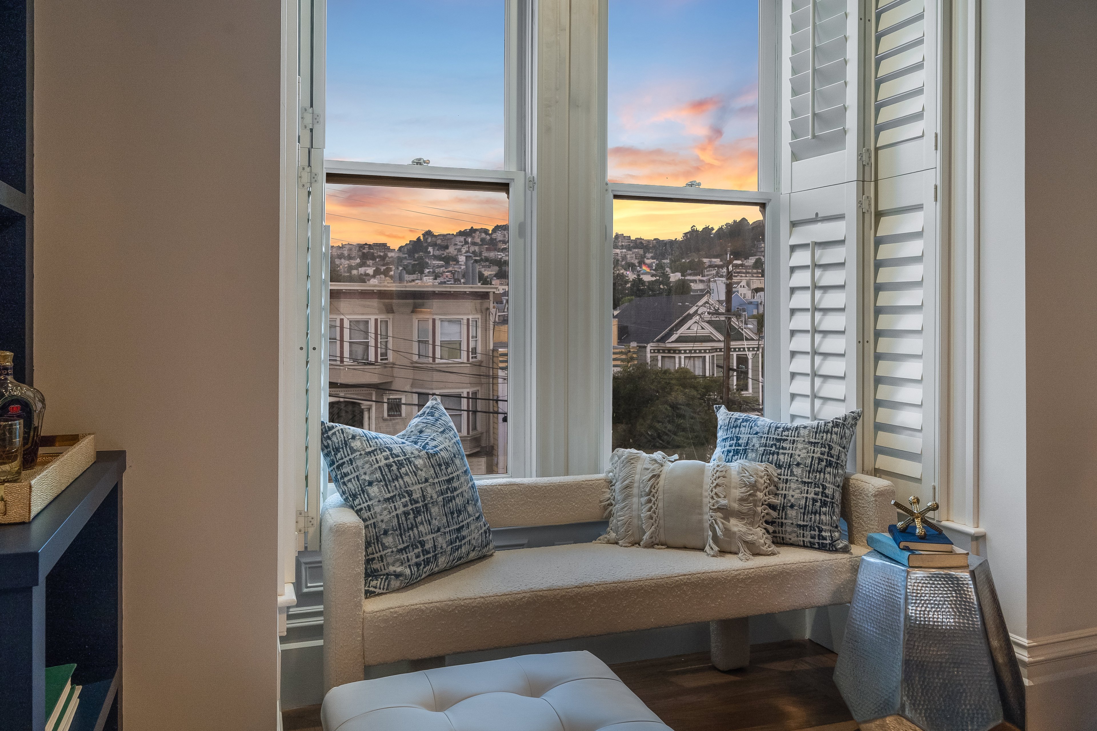 A cozy nook with a cushioned bench and decorative pillows faces a large window with white shutters, offering a view of a scenic sunset over a hilly neighborhood.