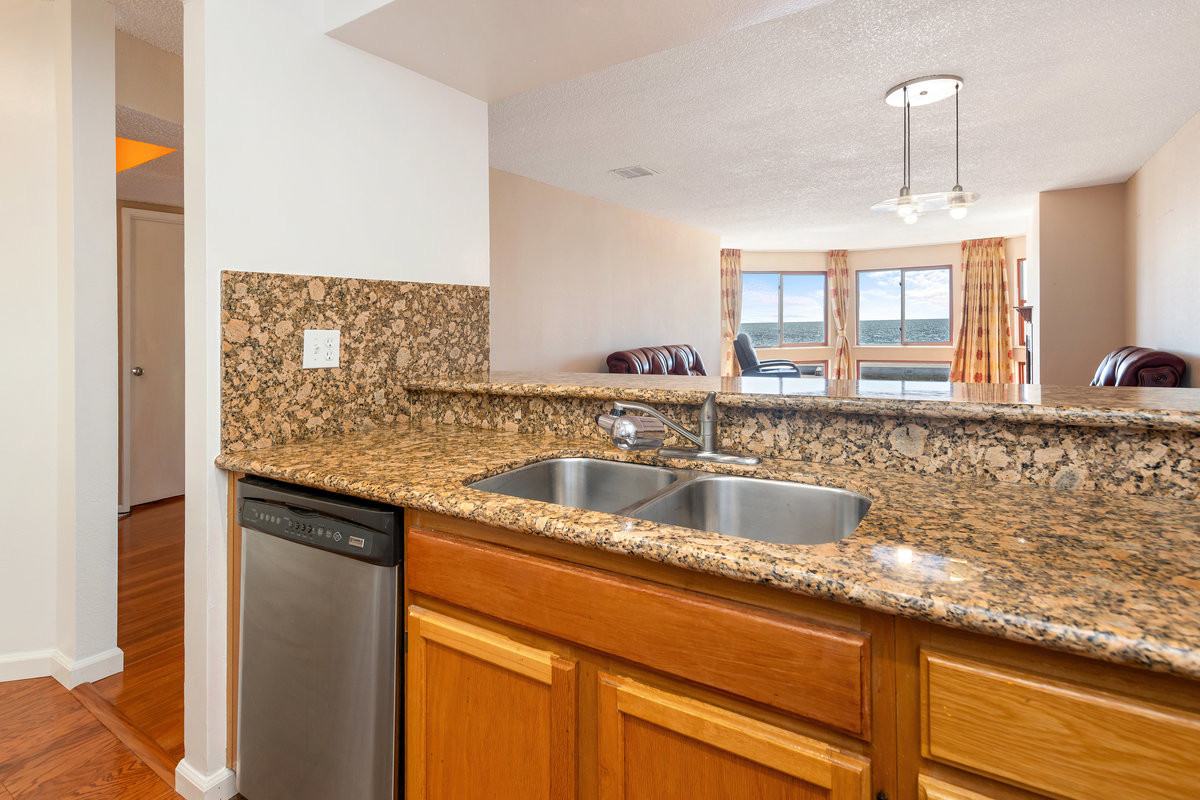 The image shows a kitchen with a granite countertop, a stainless steel double sink, and a dishwasher. It opens to a living area with large windows and a view outside.