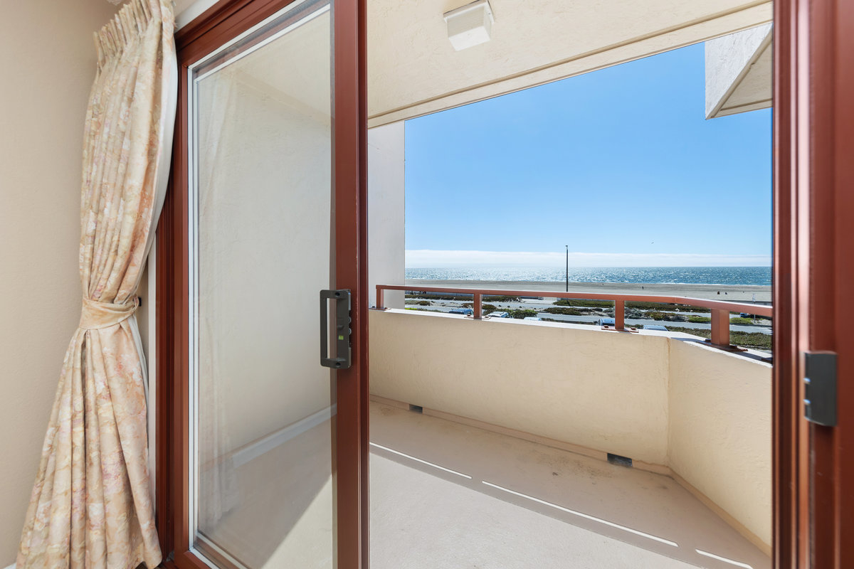 A sliding glass door, with floral curtains, opens to a small balcony with a clear view of a bright blue ocean and distant horizon.