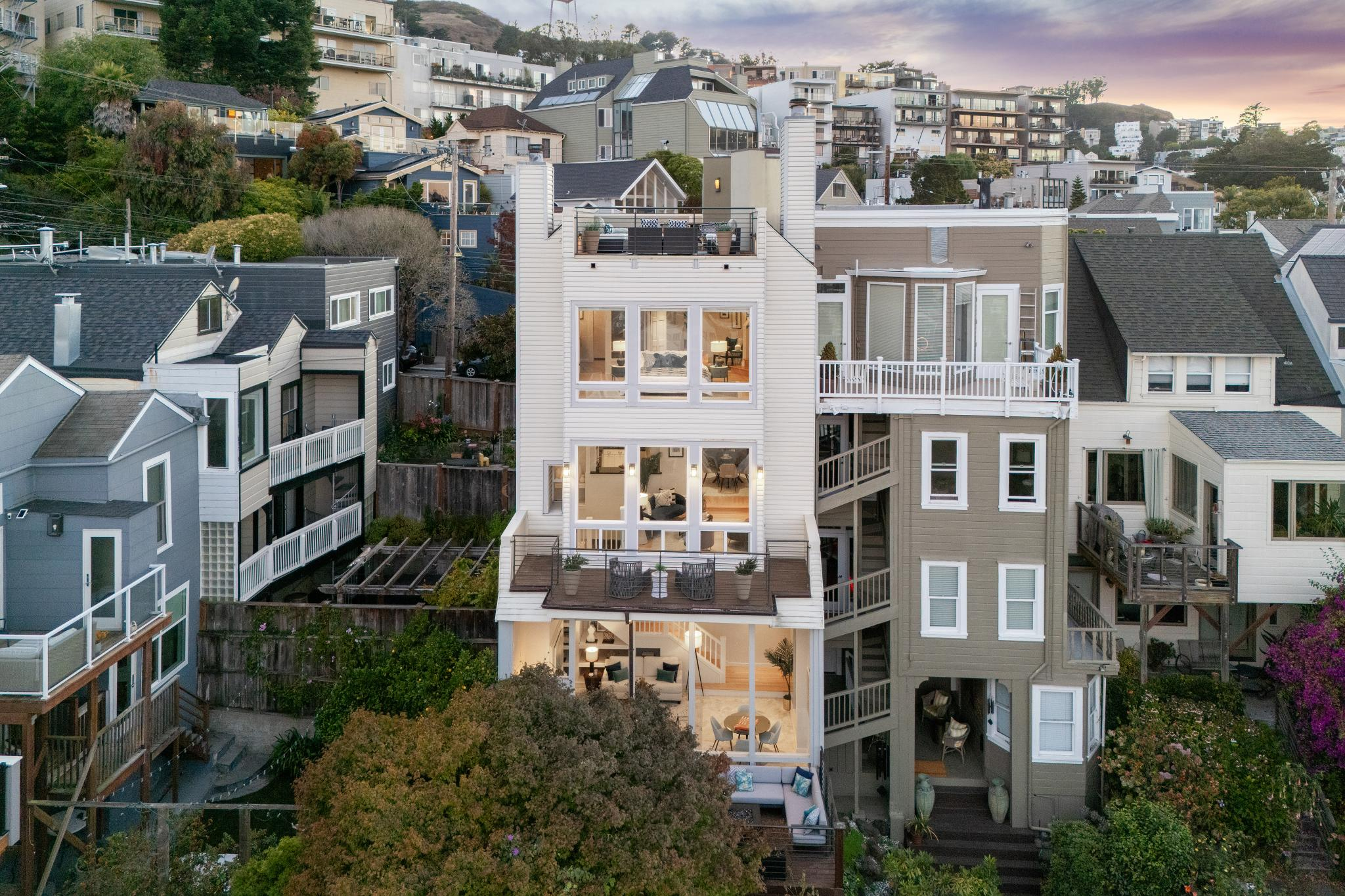 The image shows a multi-story house with large windows and balconies, set on a hillside among other similar homes. The area is lush and the sky is at sunset.