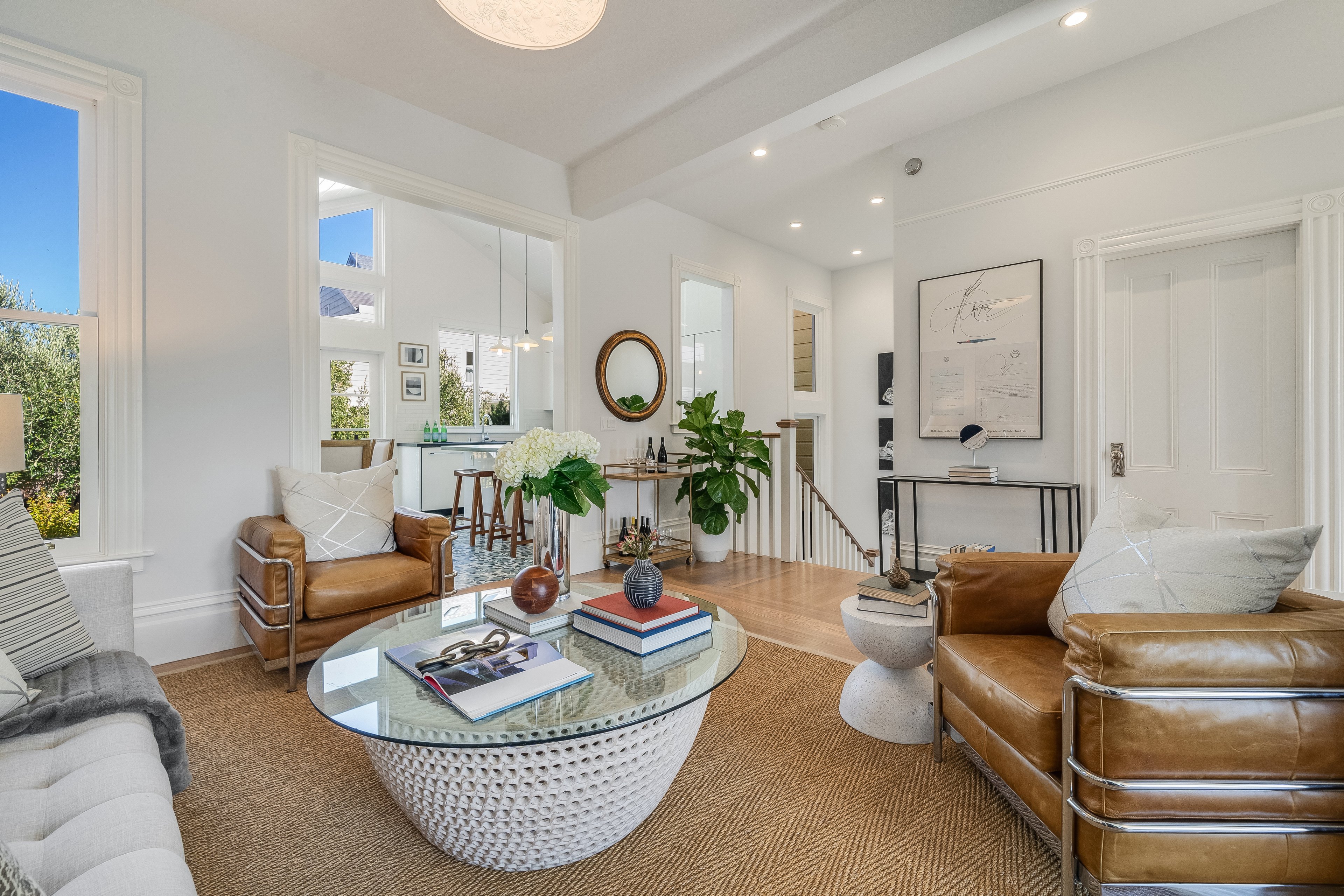 A bright living room with leather chairs, a glass coffee table, and wall art, featuring plants and modern decor, a staircase, and large windows providing natural light.