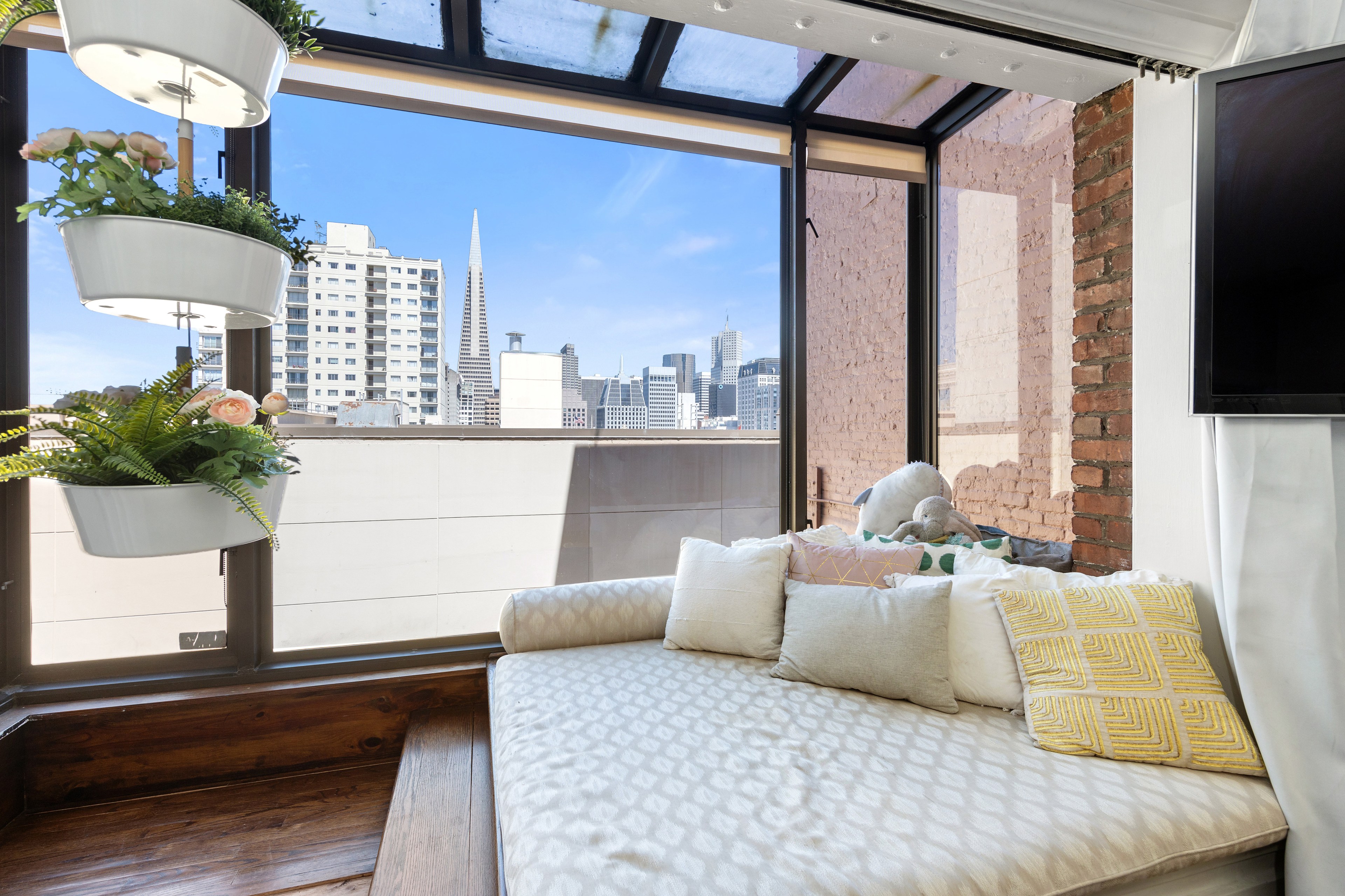 A cozy nook with a large cushioned window seat adorned with pillows, overlooking a cityscape. Hanging planters with flowers are on the left, and a TV is on the right.