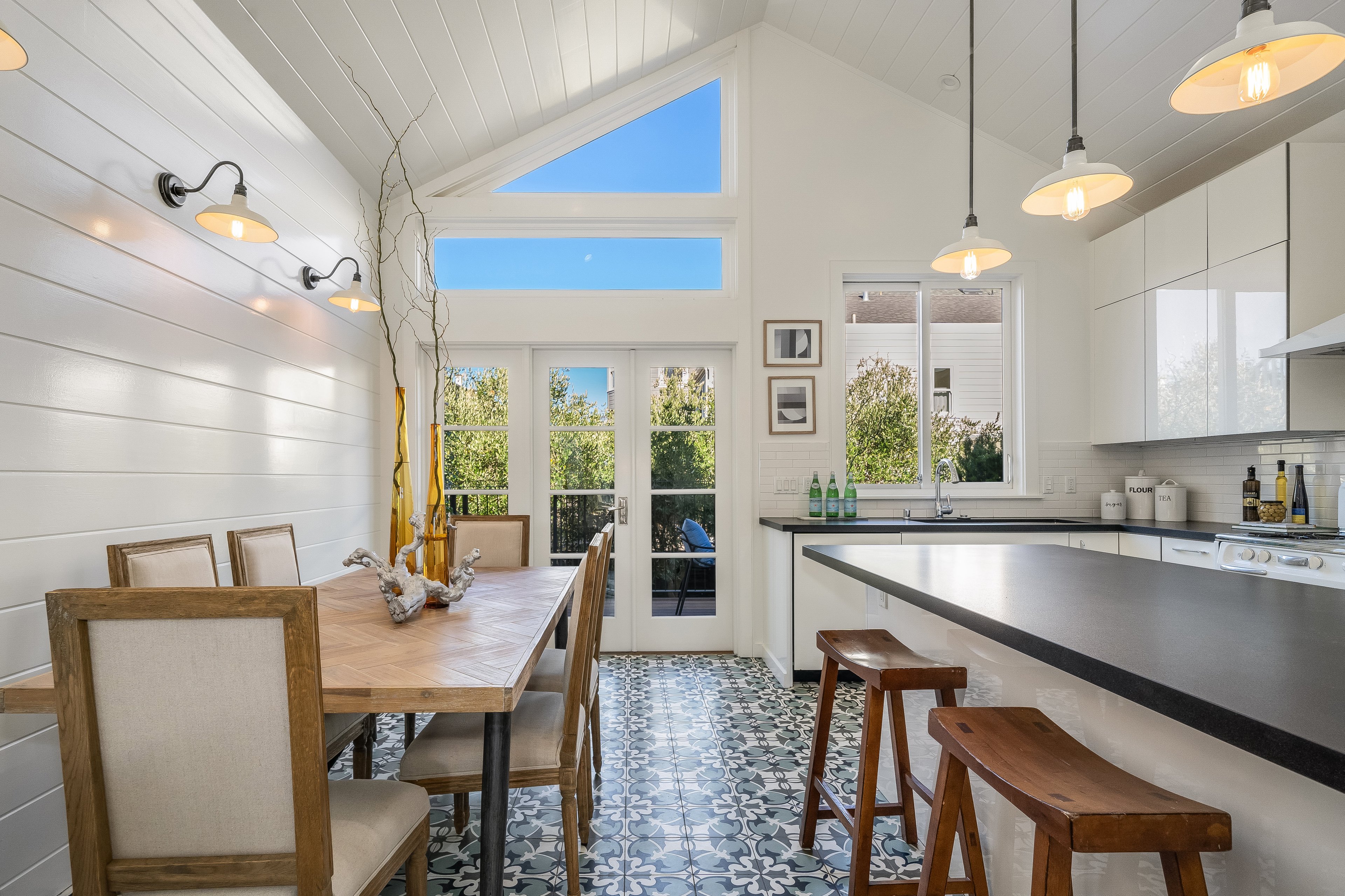 This airy kitchen features a wooden dining table, patterned floor tiles, white walls and cabinets, ample windows, pendant lights, and a black countertop island.