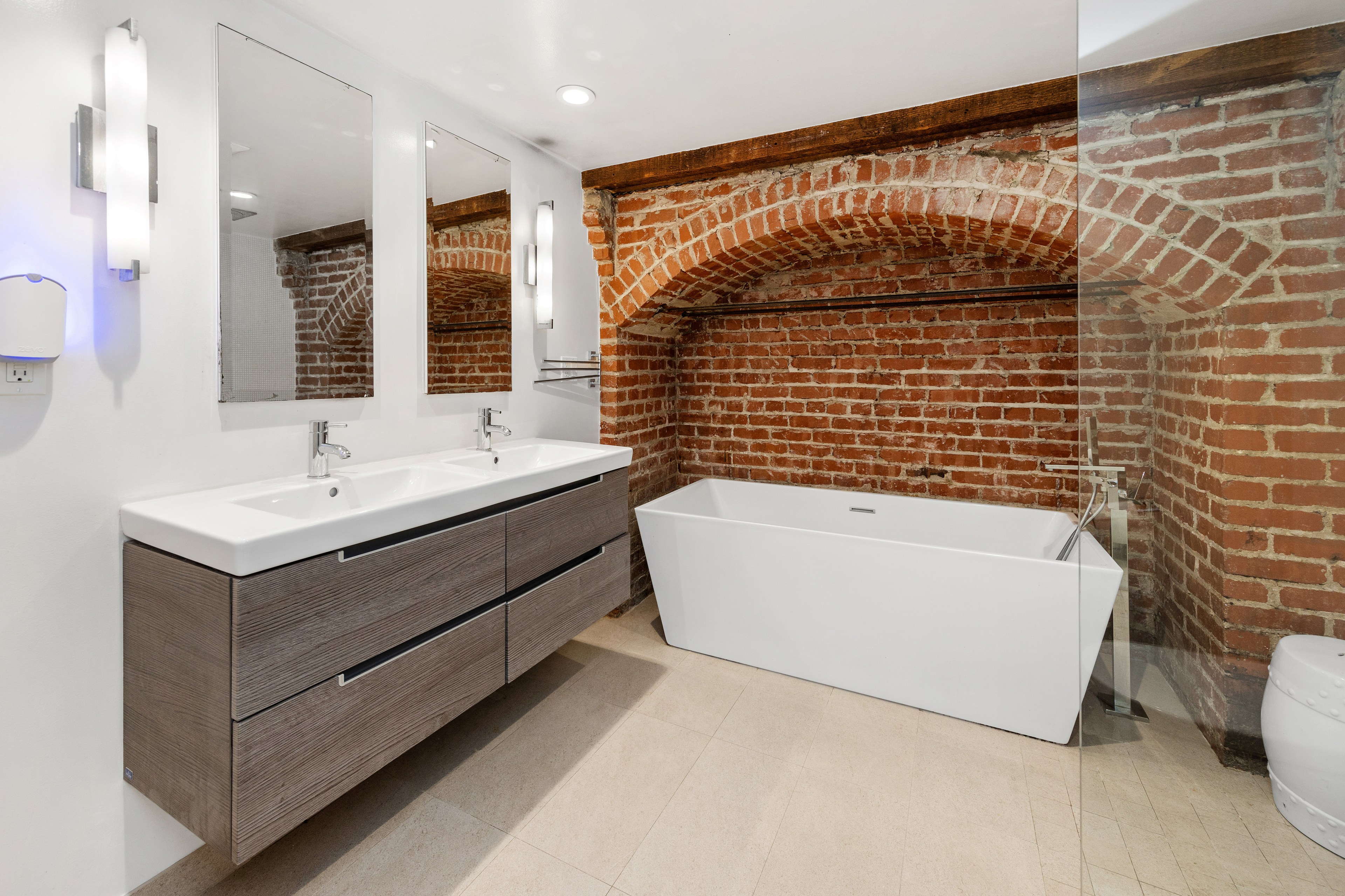 The bathroom features a double sink vanity with two mirrors, a modern white bathtub against a brick wall, and beige tile flooring.