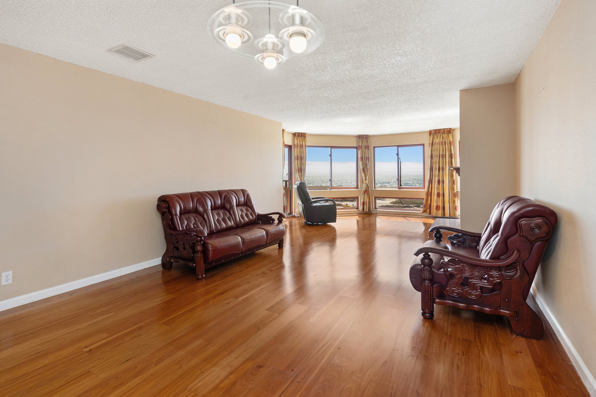 A spacious, hardwood-floored room features two leather sofas, a recliner by large windows with panoramic views, and a modern chandelier hanging from the ceiling.