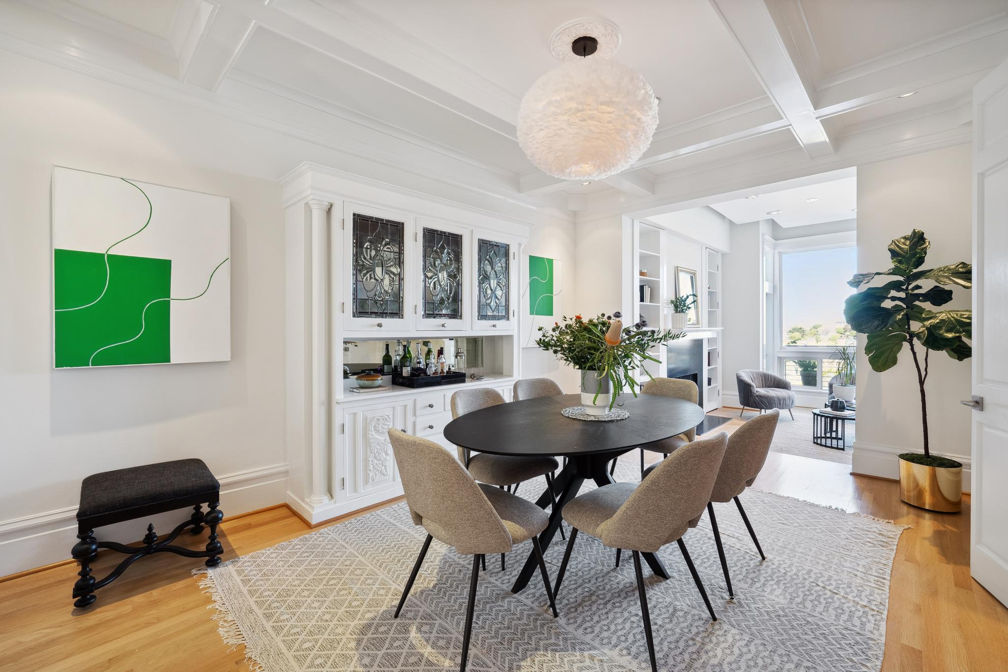 A modern dining room features a black oval table surrounded by six gray chairs, a large leafy plant, white cabinets with glass doors, abstract art, and ample natural light.