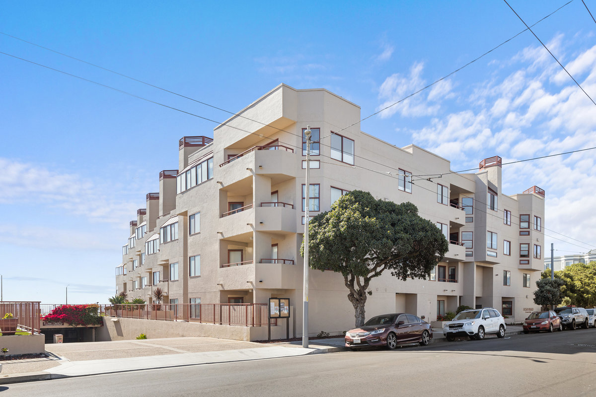 The image shows a modern, multi-story apartment complex on a sunny day, with cars parked along the street and a large tree in front.