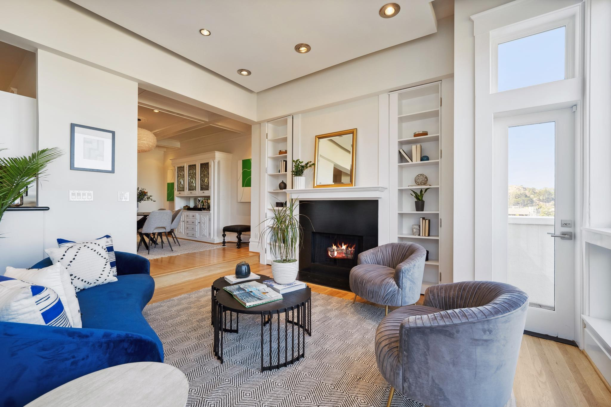 A cozy living room with a blue couch, two gray armchairs, and a coffee table in front of a fireplace. Shelves flank the fireplace, a dining area is seen in the background.