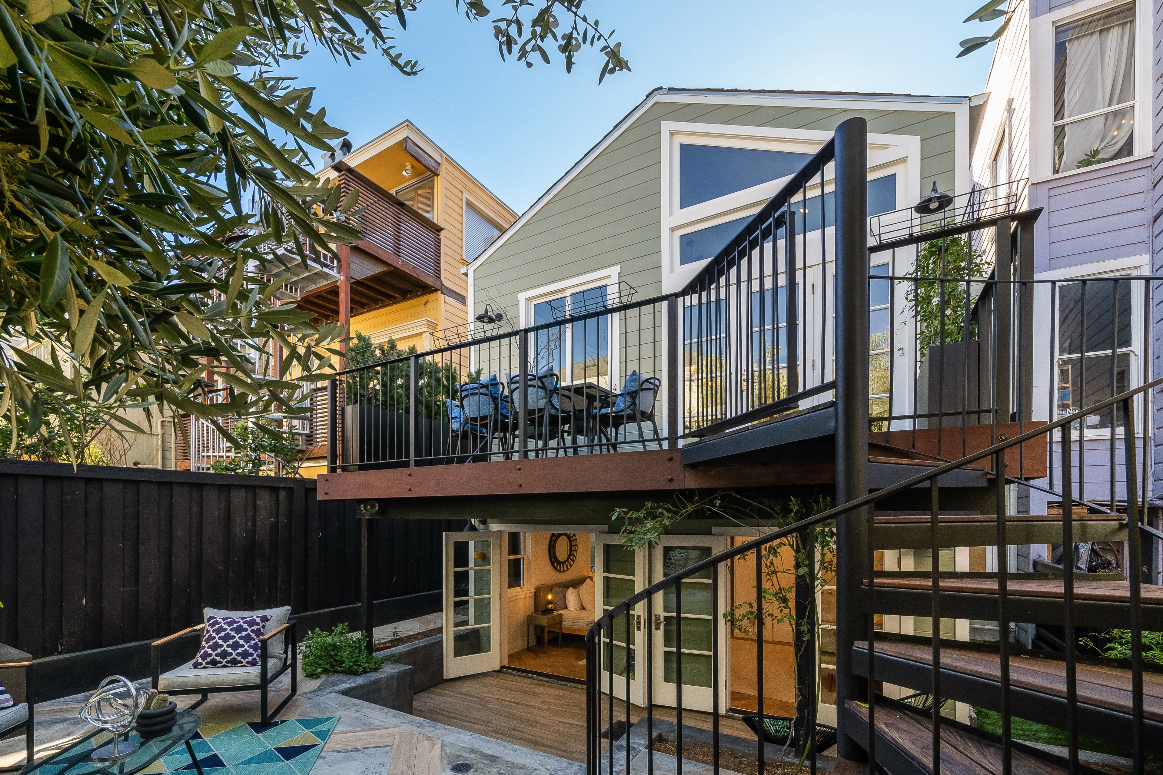 A two-level backyard with a spiral staircase features a ground patio and an upper deck with outdoor seating attached to a green house, surrounded by neighboring homes.