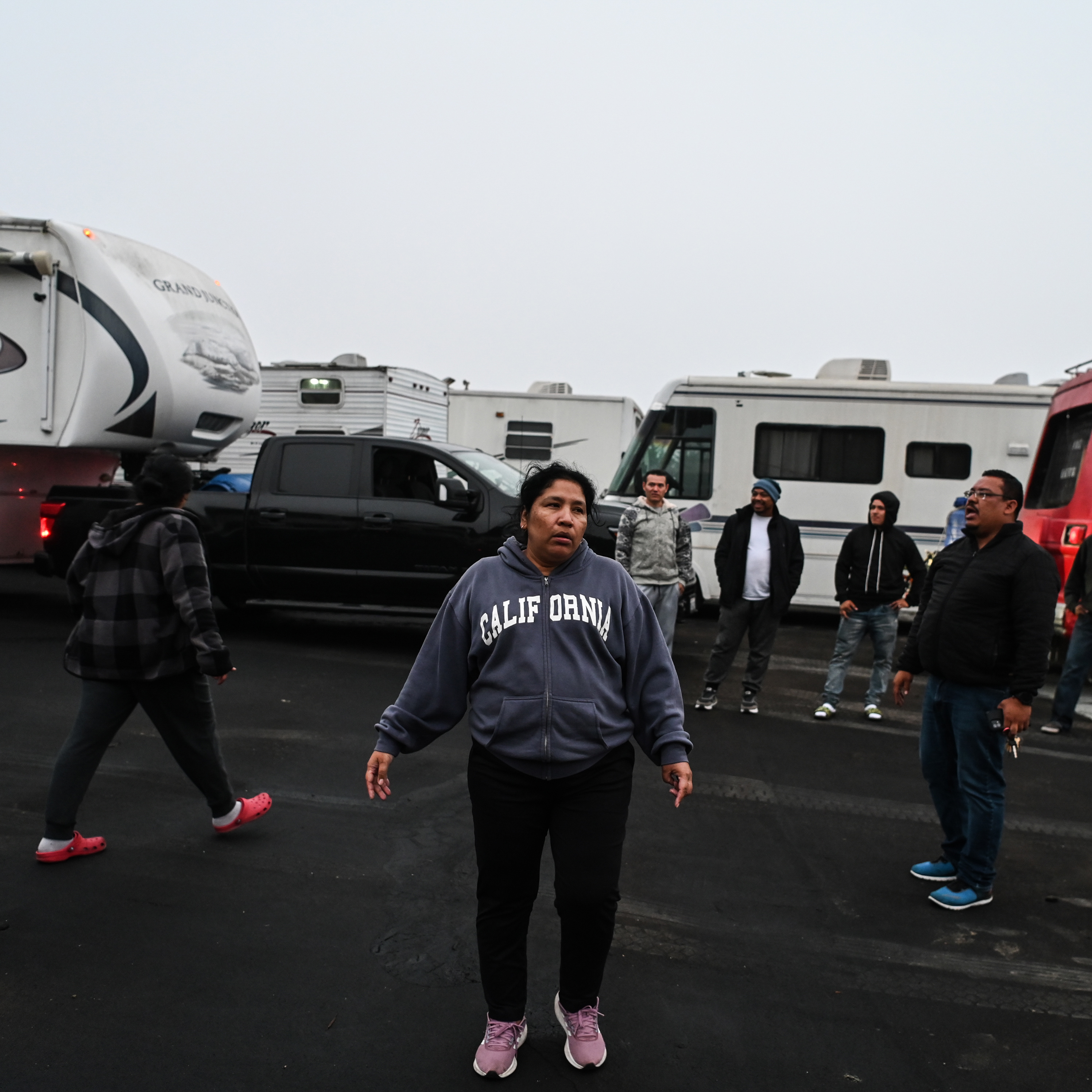 A woman in a "California" hoodie stands on a paved lot with RVs and trucks, surrounded by several people in casual clothing.