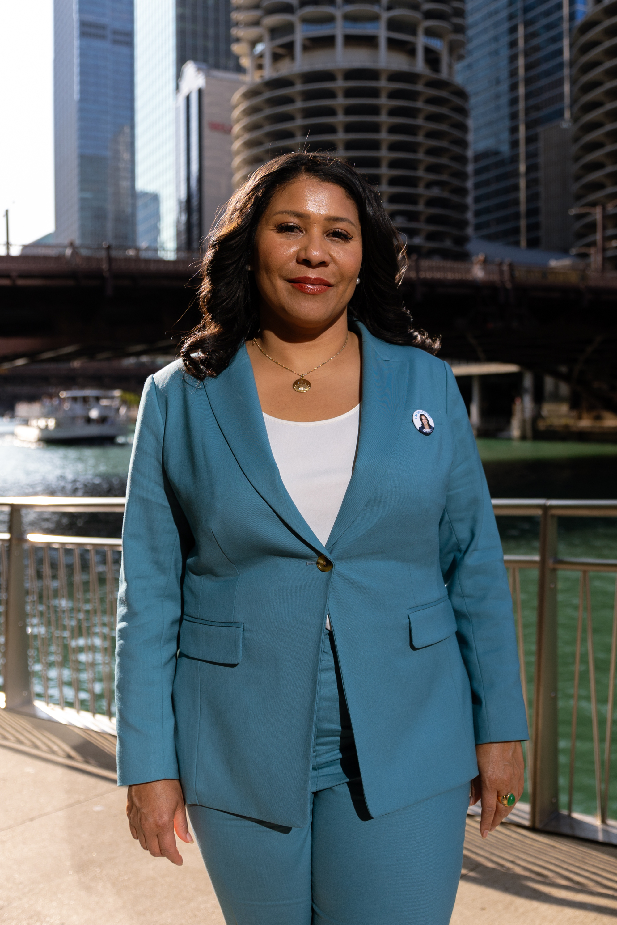 A woman in a blue suit stands smiling by a river, with modern high-rise buildings and a bridge in the background. She wears a white top and a gold necklace.