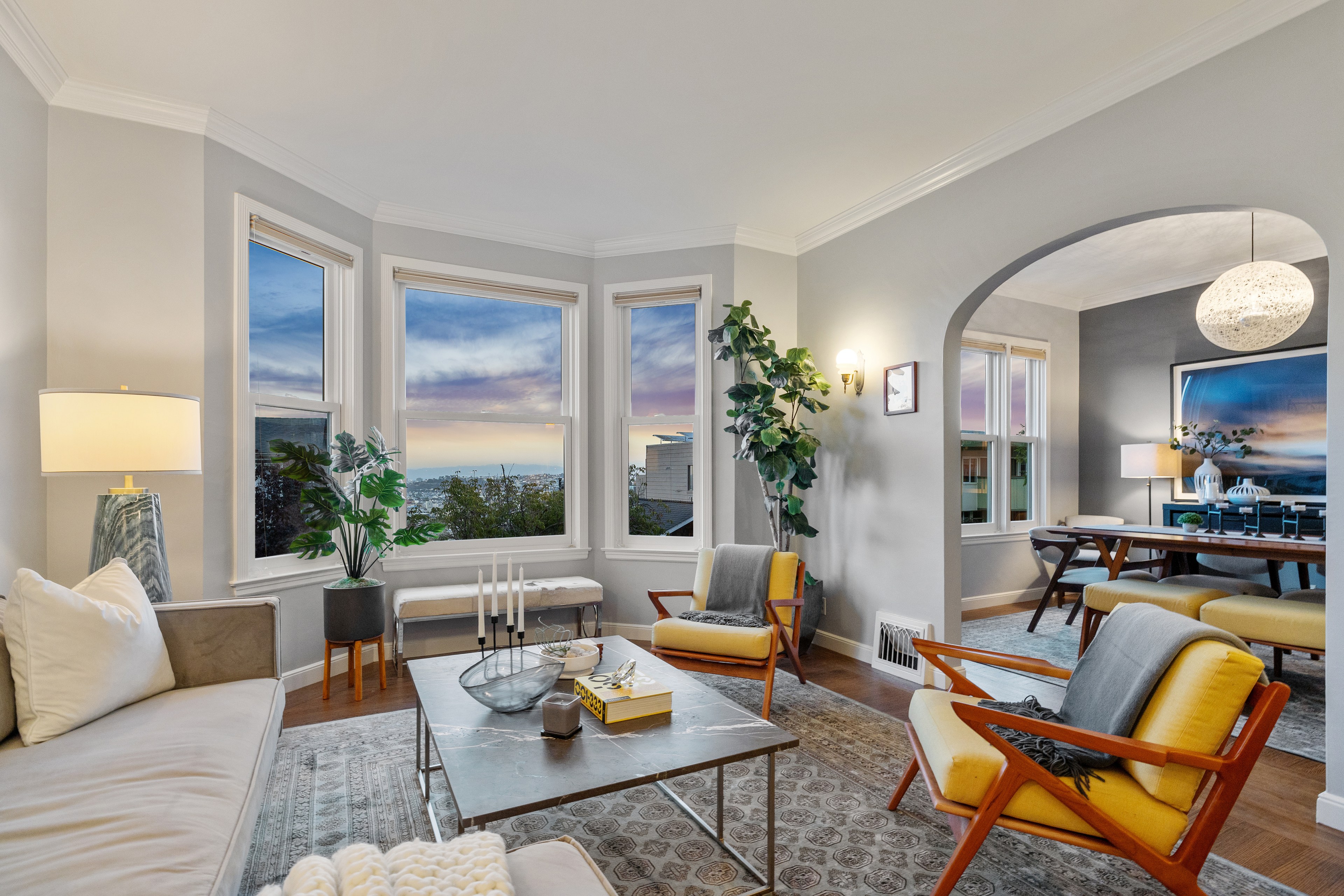 A stylish living room with a couch, two yellow chairs, a coffee table, plants, and large windows with a sunset view, adjoining a dining area through an arched opening.