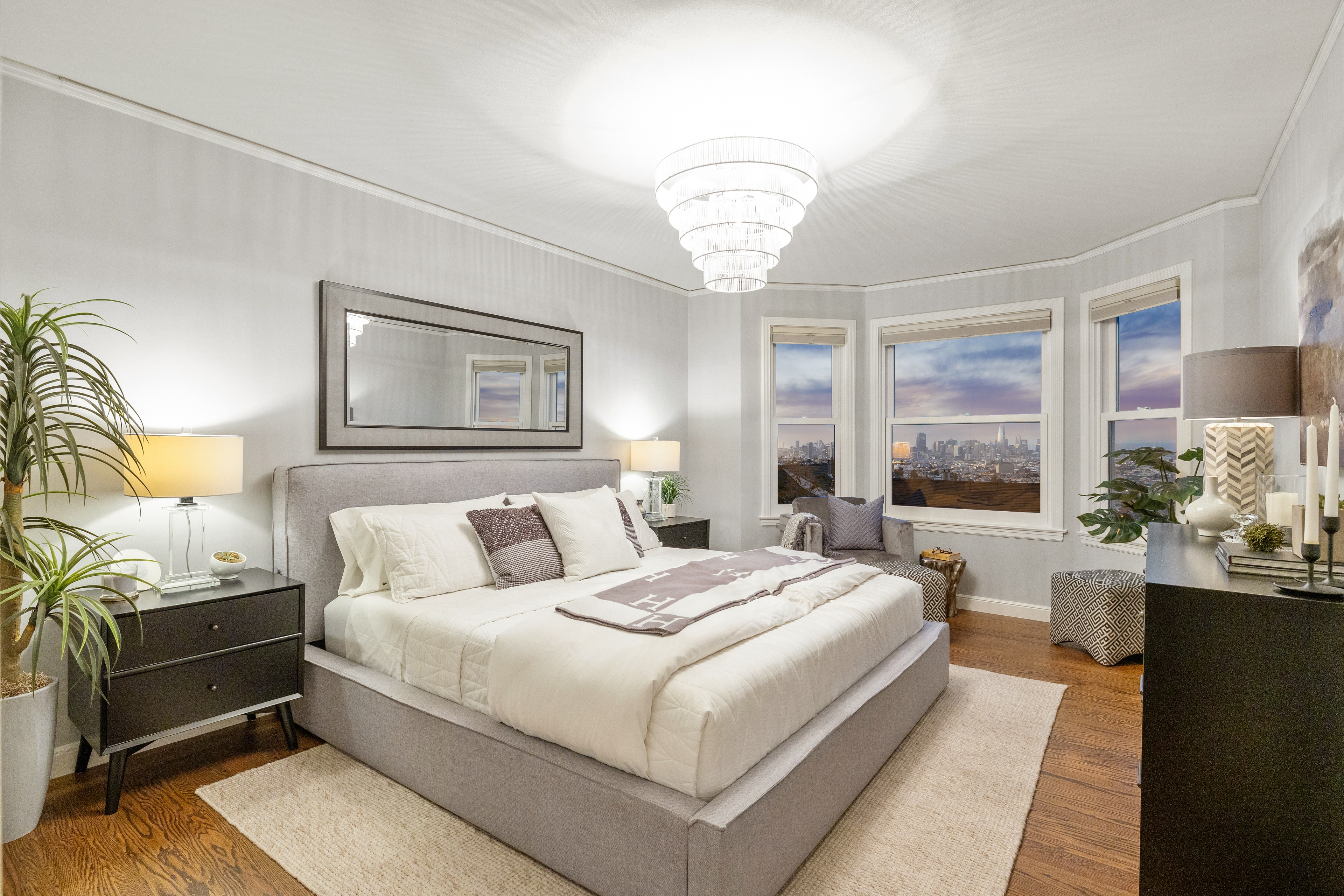 A cozy bedroom with a large bed, elegant chandeliers, and three windows showcasing a city skyline view. It contains modern furniture, lamps, and decorative plants.