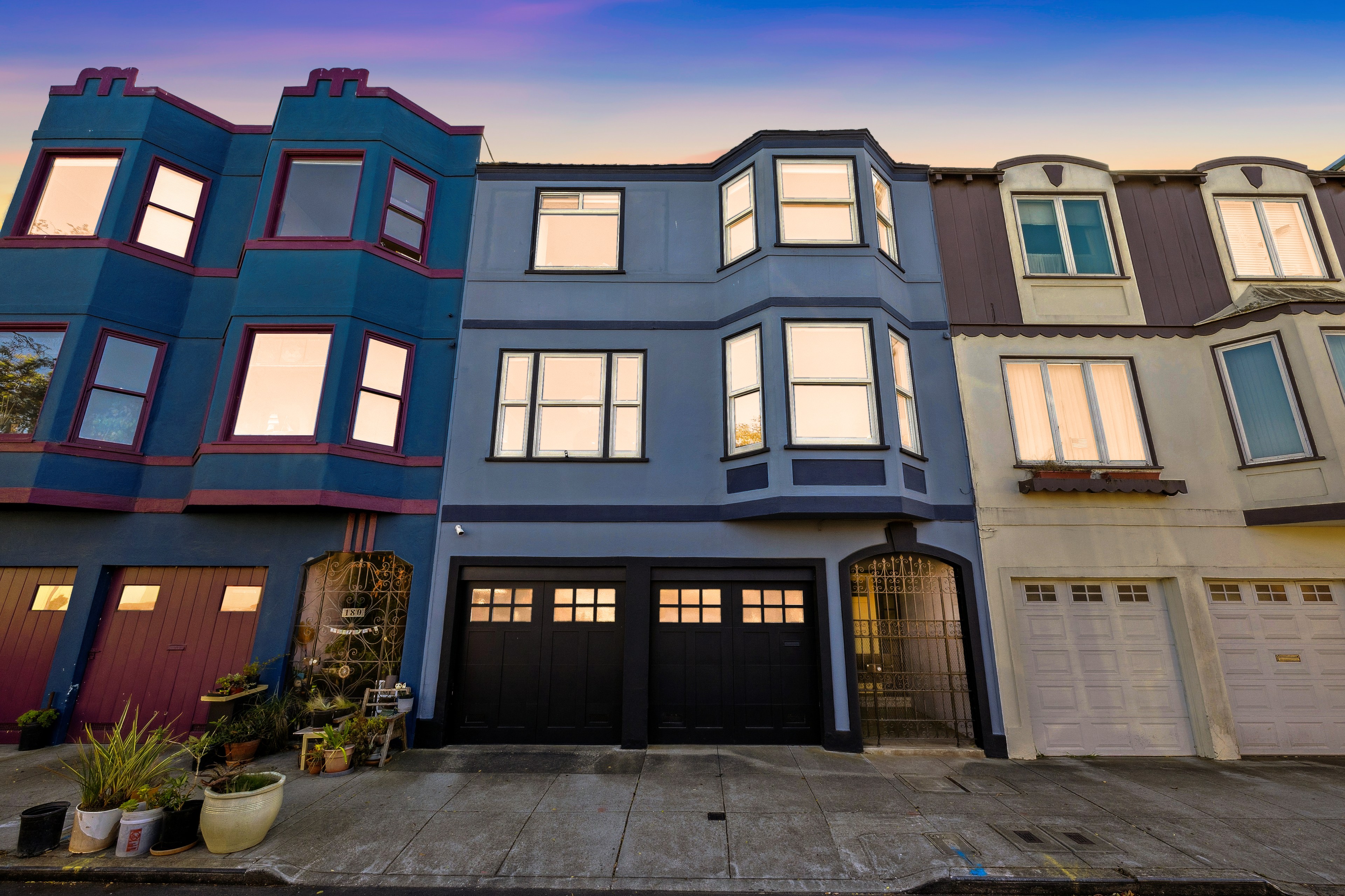 The image shows a row of colorful townhouses with bay windows. One house is blue with red-trimmed windows, while the others are blue and white. The sky is a gradient at dusk.