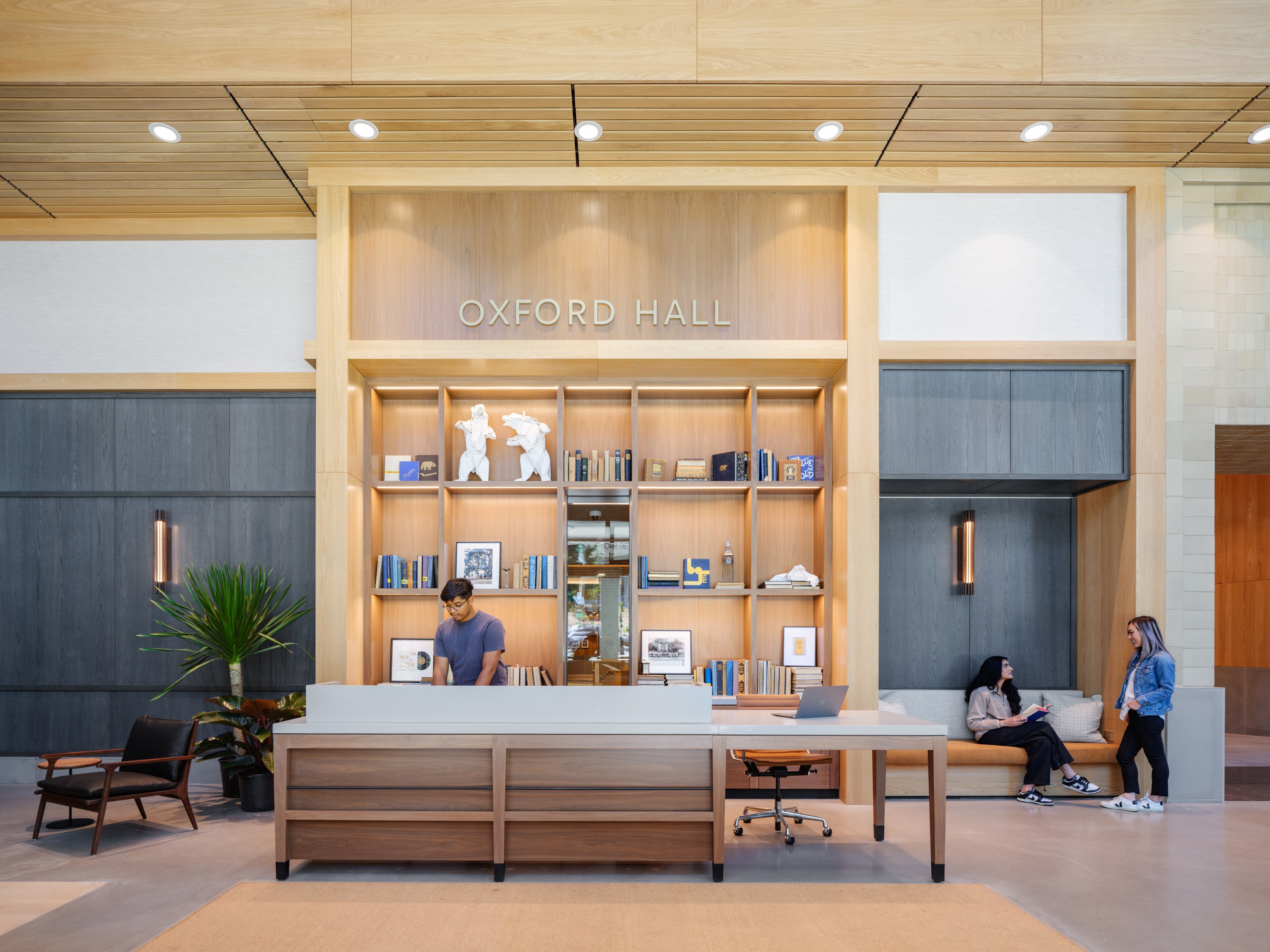 The image shows a modern reception area with a man standing at a desk beneath a sign reading &quot;Oxford Hall.&quot; Two women are seated and standing on the right.