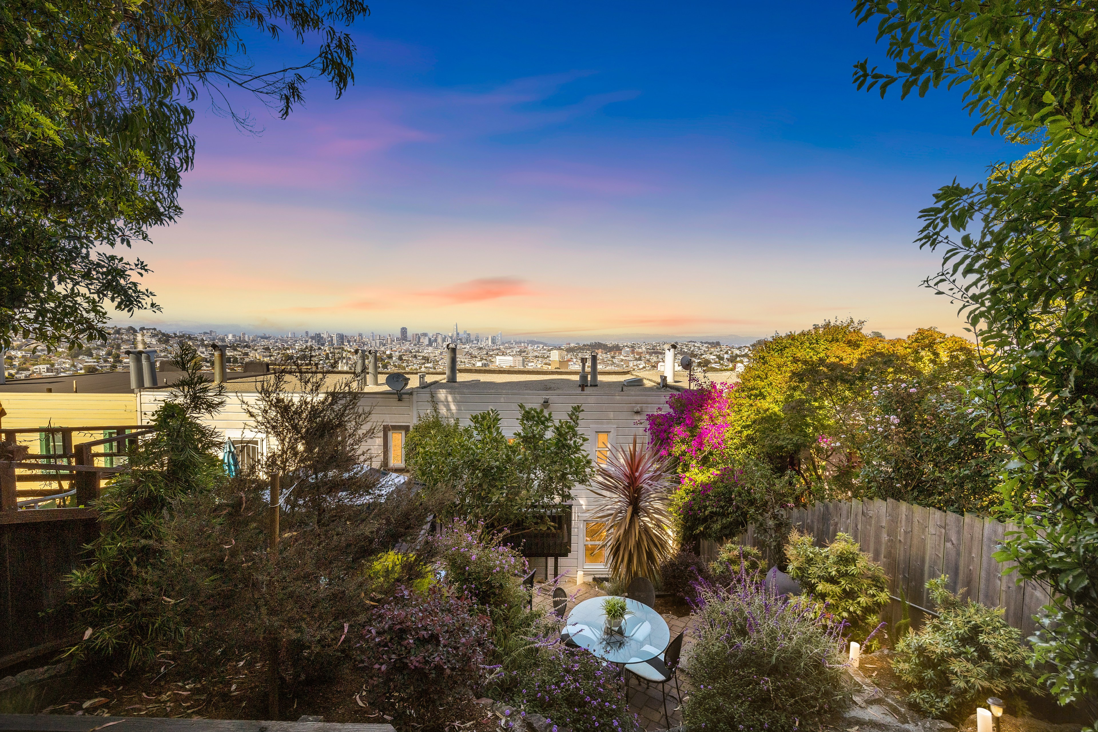 A lush garden with various plants surrounds a patio set. The scene overlooks a cityscape at sunset, with rooftops and distant skyscrapers under a blue and pink sky.