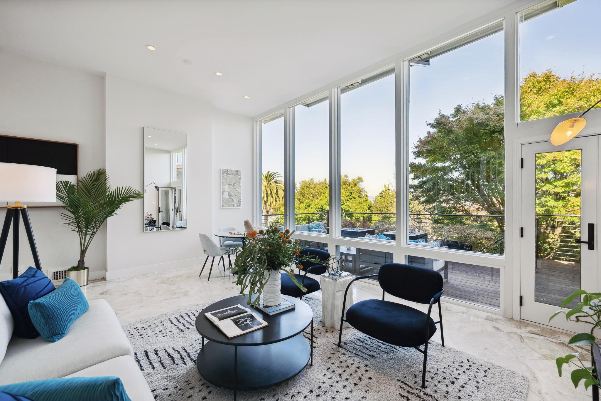 A bright, modern living room features floor-to-ceiling windows, white walls, a white sofa with blue cushions, black chairs, a round coffee table, and a lush outdoor view.