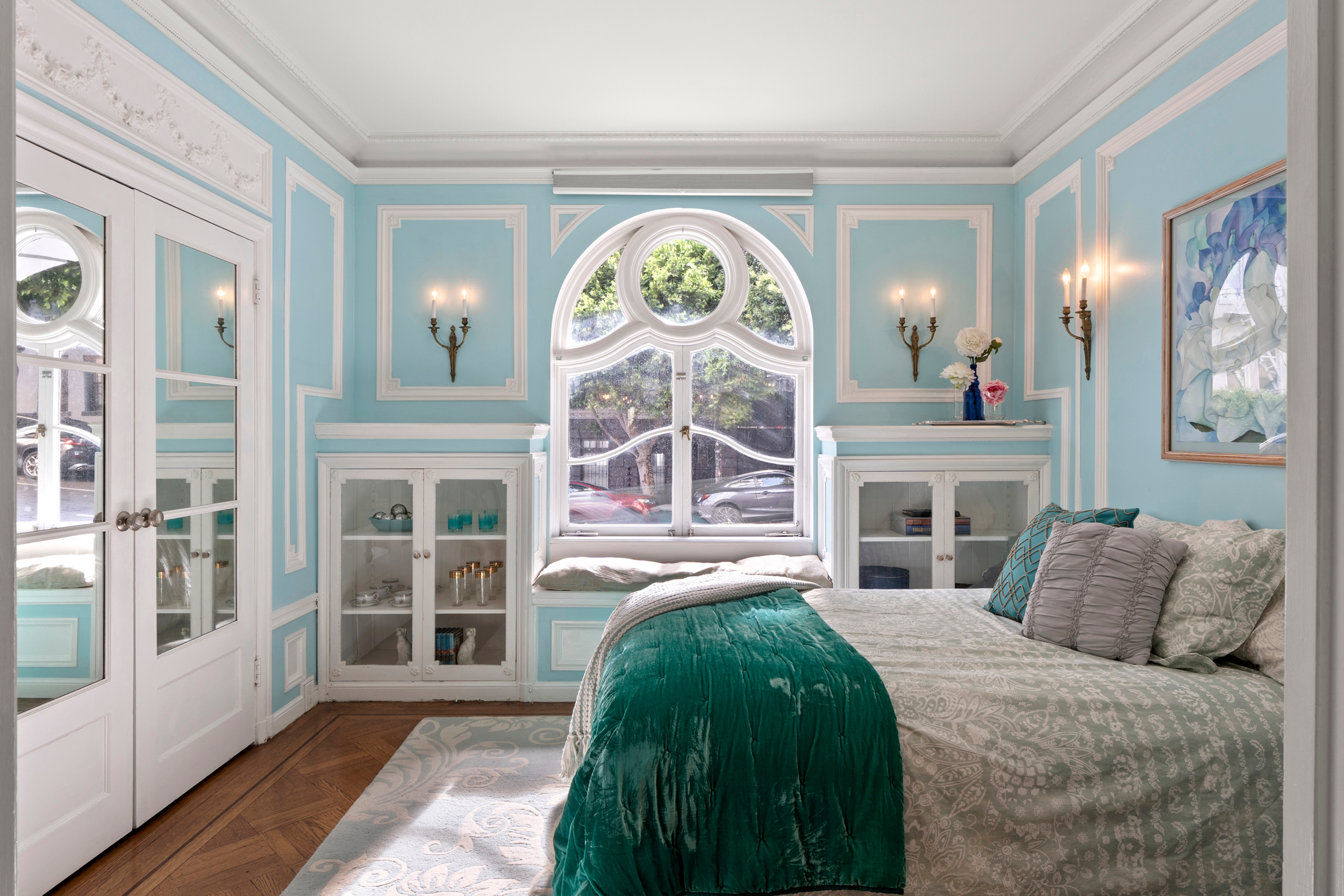 This bedroom features aqua walls with white trim, ornate sconces, a large bed with green and gray bedding, and a round window flanked by mirrored cabinets.