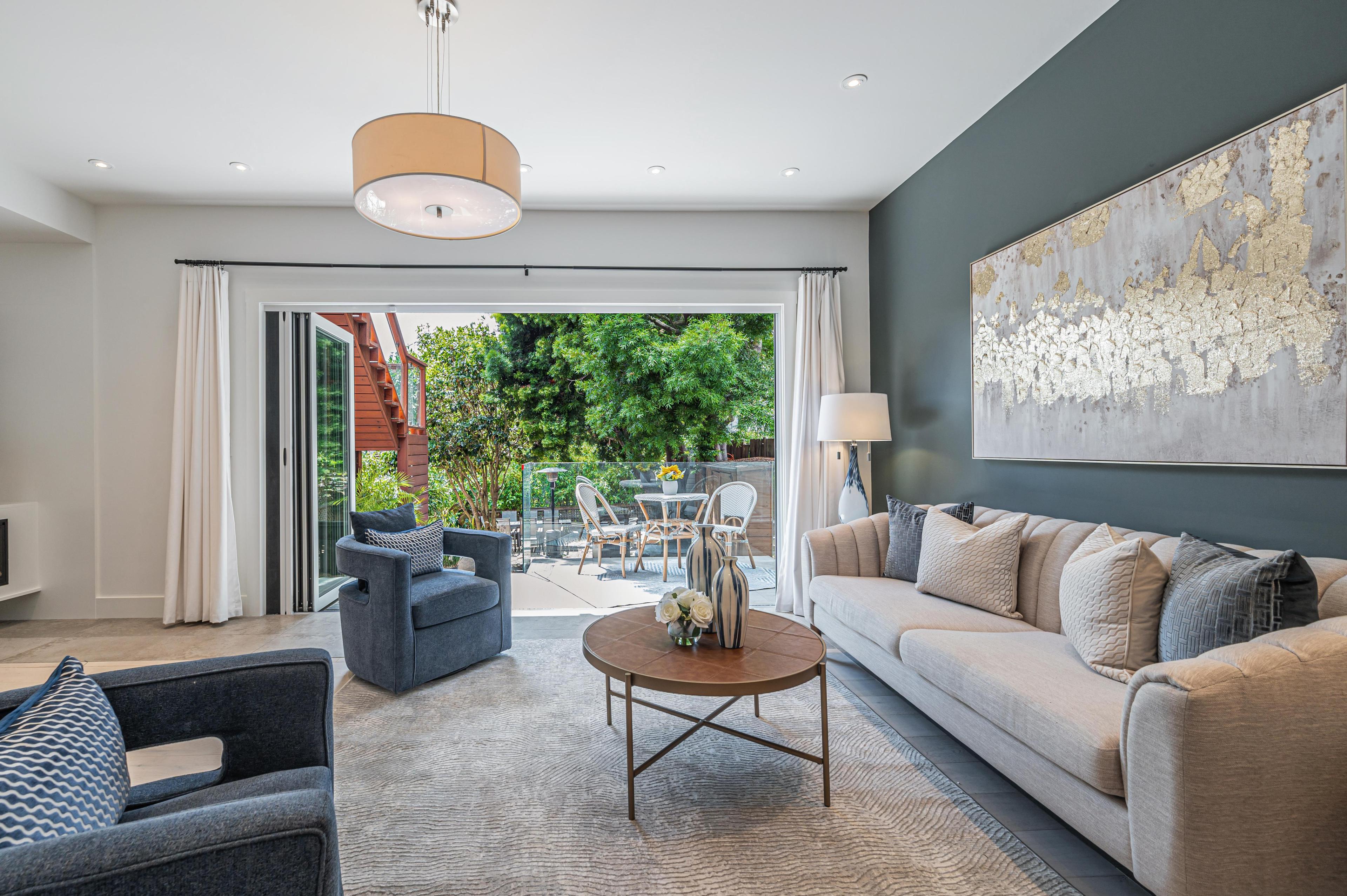 A modern living room with a light beige sofa, gray armchairs, a round wooden coffee table, and large artwork on a gray accent wall. Glass doors lead to a patio.