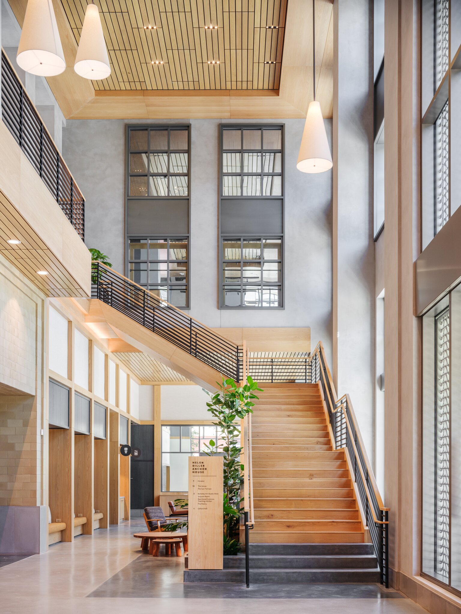 The image shows a modern, spacious interior with high ceilings, wooden stairs, large windows, and stylish pendant lights. Plants and wooden furniture add a warm touch.