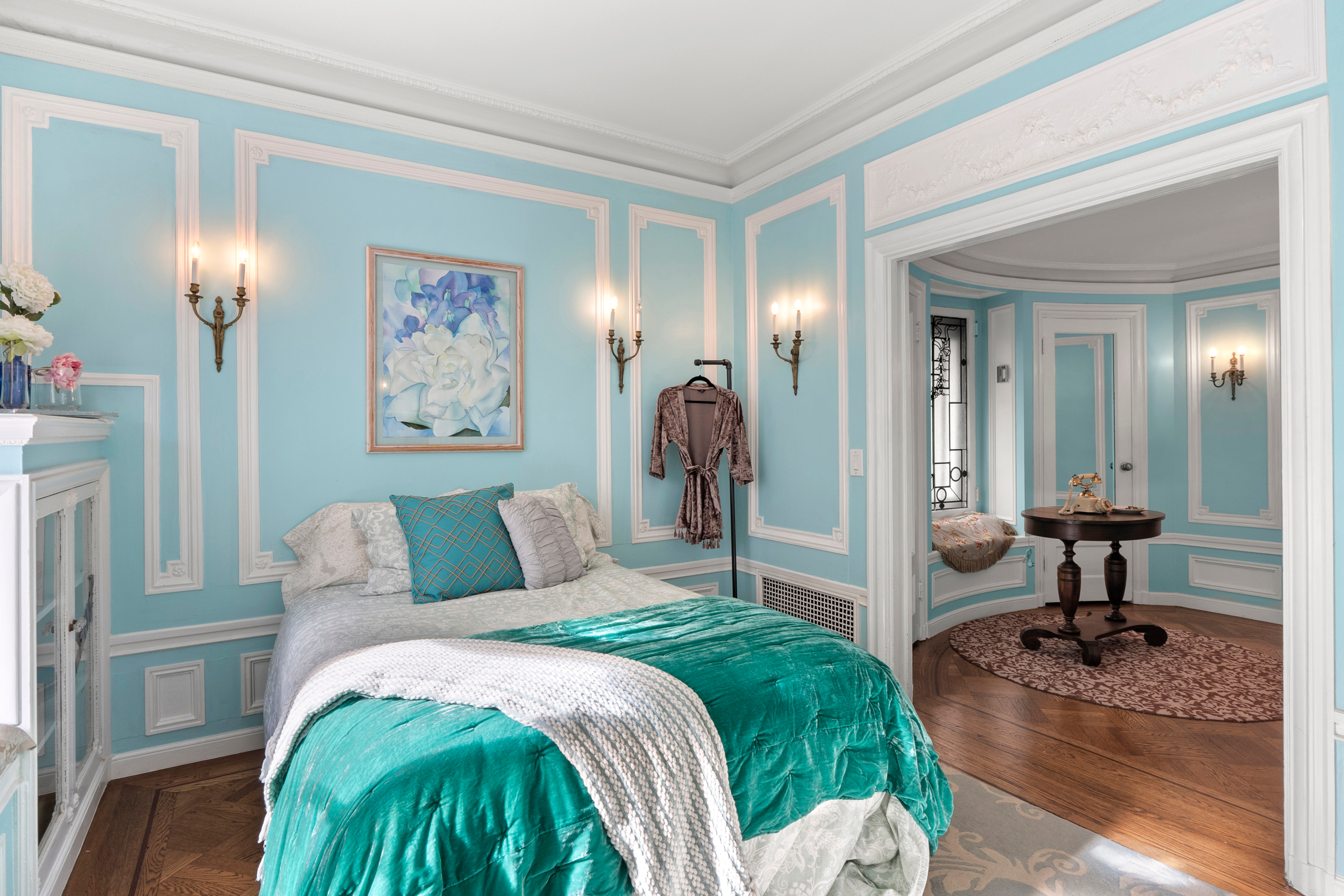 This elegant bedroom features light blue walls with white trim, a neatly made bed with teal and white bedding, wall sconces, and a hallway leading to a circular room.