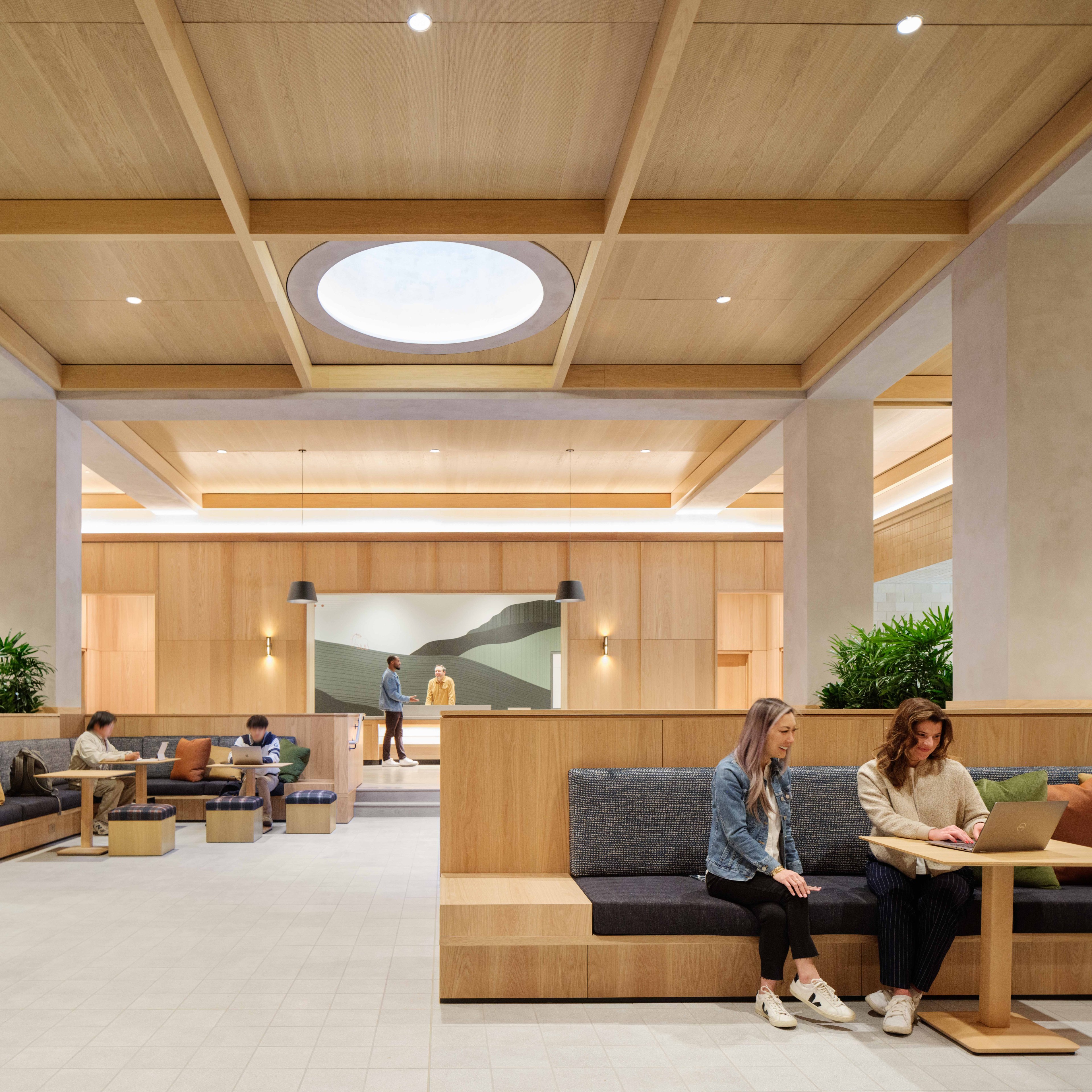 A modern lounge space with wooden interiors features people sitting, working on laptops, and talking. Large plants, comfortable seating, and a circular skylight are present.