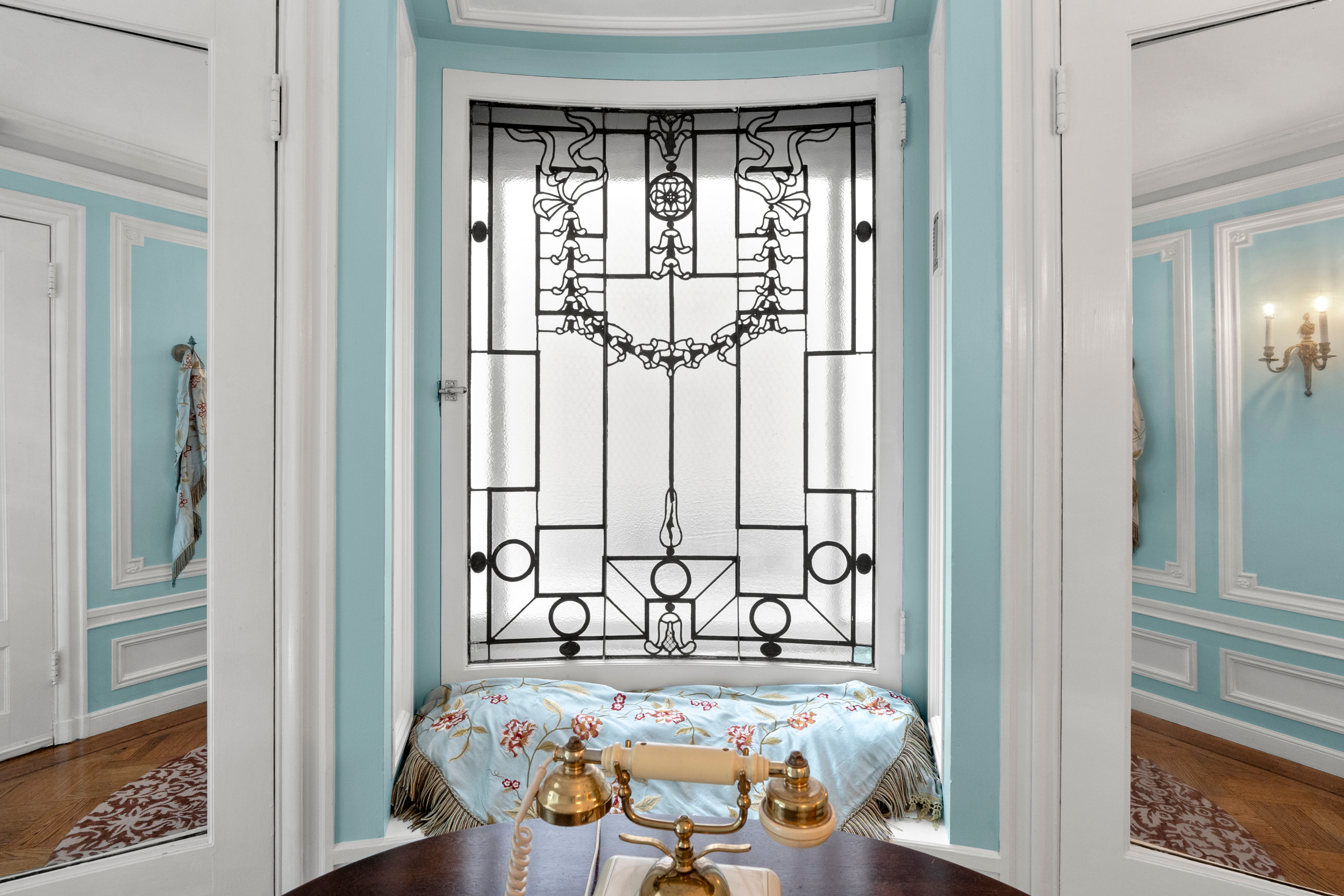 A decorative stained glass window with black geometric and floral designs is framed by light blue walls and white trim. Beneath the window is a cushioned seat with a floral cover.