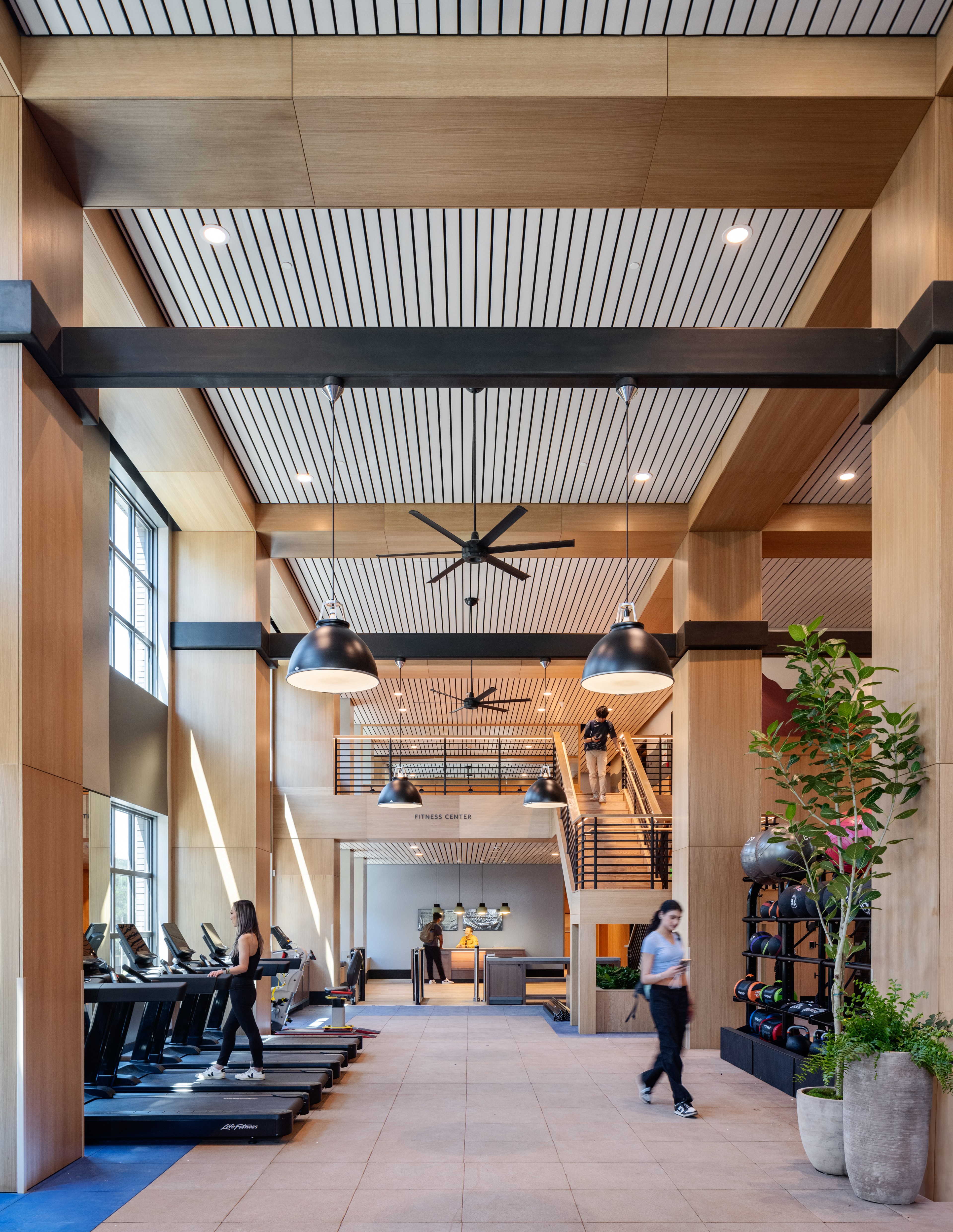 The image shows a spacious gym with high ceilings, wooden beams, exercise equipment, a person on a treadmill, two people walking, and a staircase leading to &quot;Fitness Center.&quot;